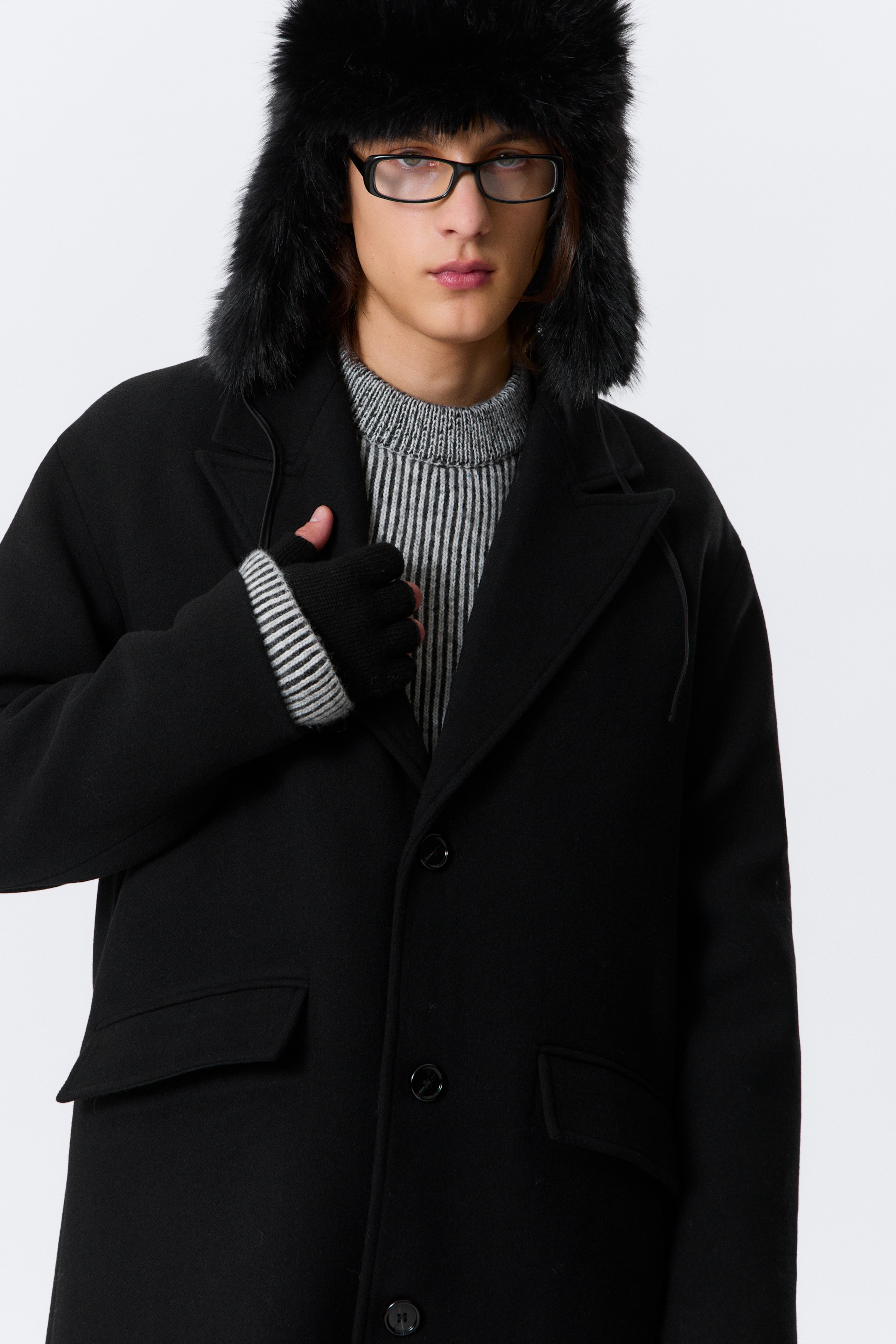 View larger image: A black coat is being shown from the front, angled slightly to the right, featuring notch lapels, a three-button front closure, and two flap pockets on the lower front. A striped, ribbed sweater is being worn underneath, with black fingerless gloves visible at the wrists.