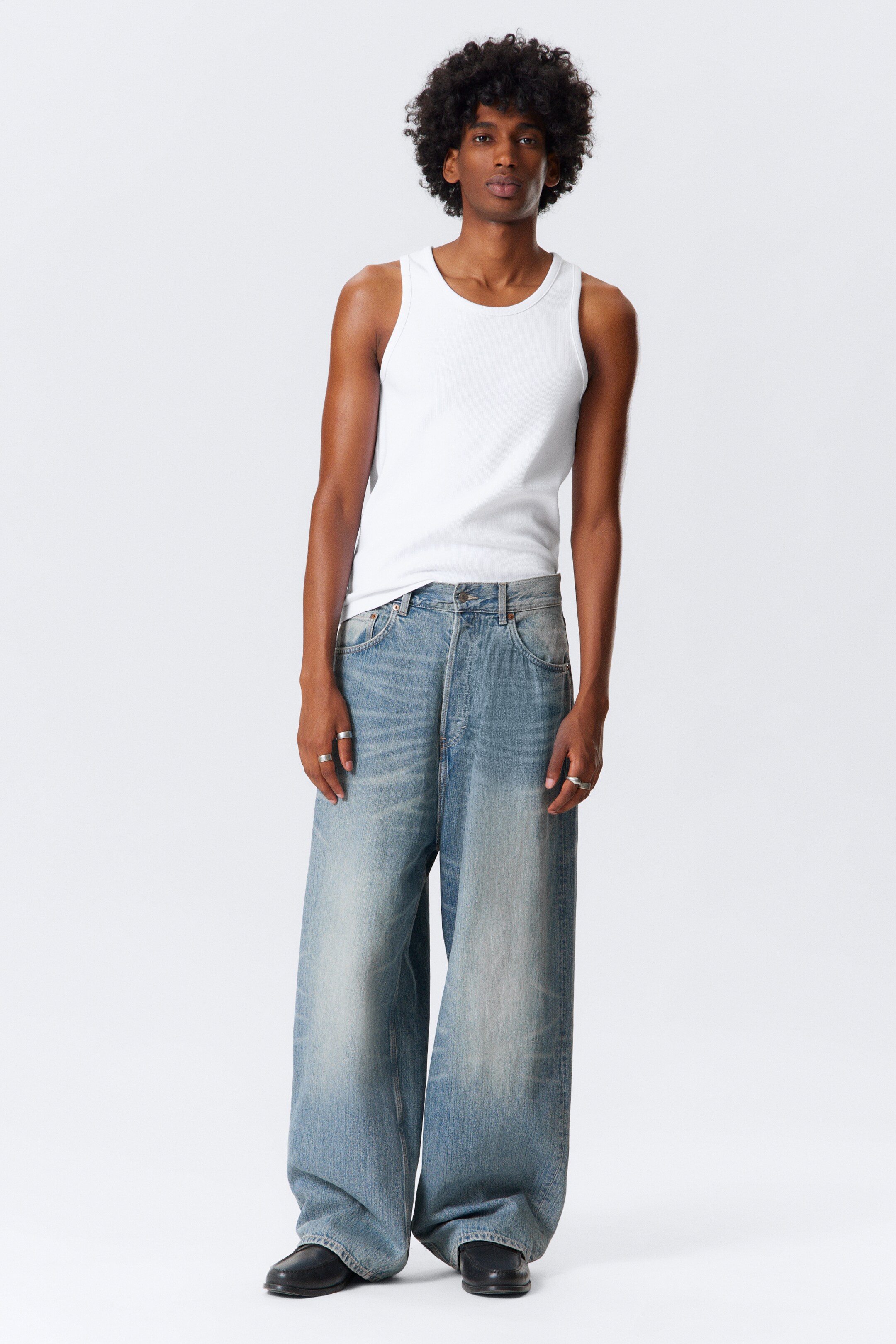 Grotere afbeelding bekijken: Een witte geribbelde tanktop wordt gedragen, met een ronde hals en een nauwsluitende pasvorm. Het wordt gecombineerd met lichtgewassen, wijde jeans die zijn ontworpen met een ontspannen, baggy silhouet.