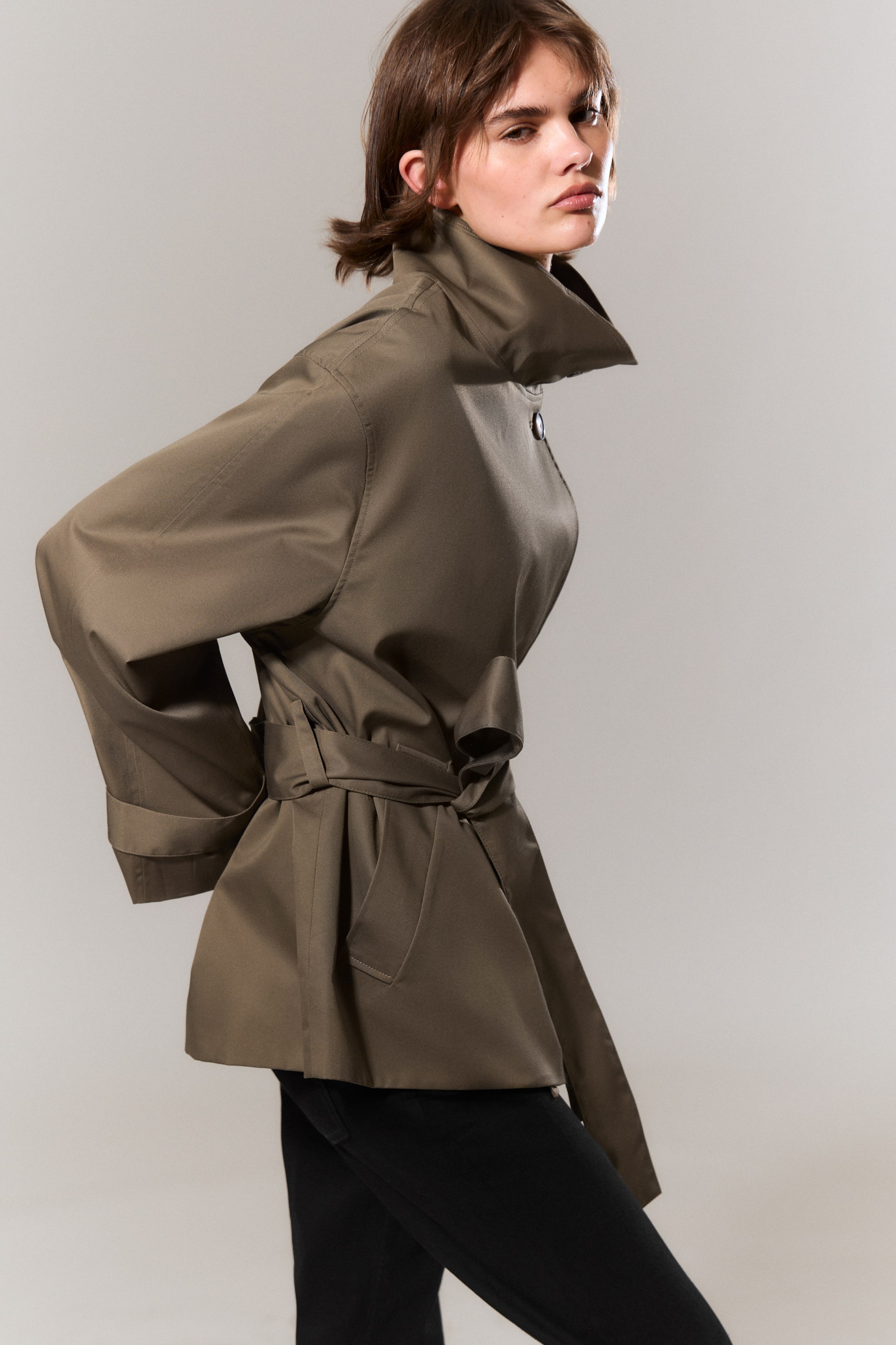 View larger image: A khaki jacket, viewed from a side angle, features a high standing collar with a single button fastening at the neck. It cinches at the waist with a self-fabric tie-belt, creating a flared hem, and showcases wide sleeves with folded cuffs and a visible patch pocket on the lower front.