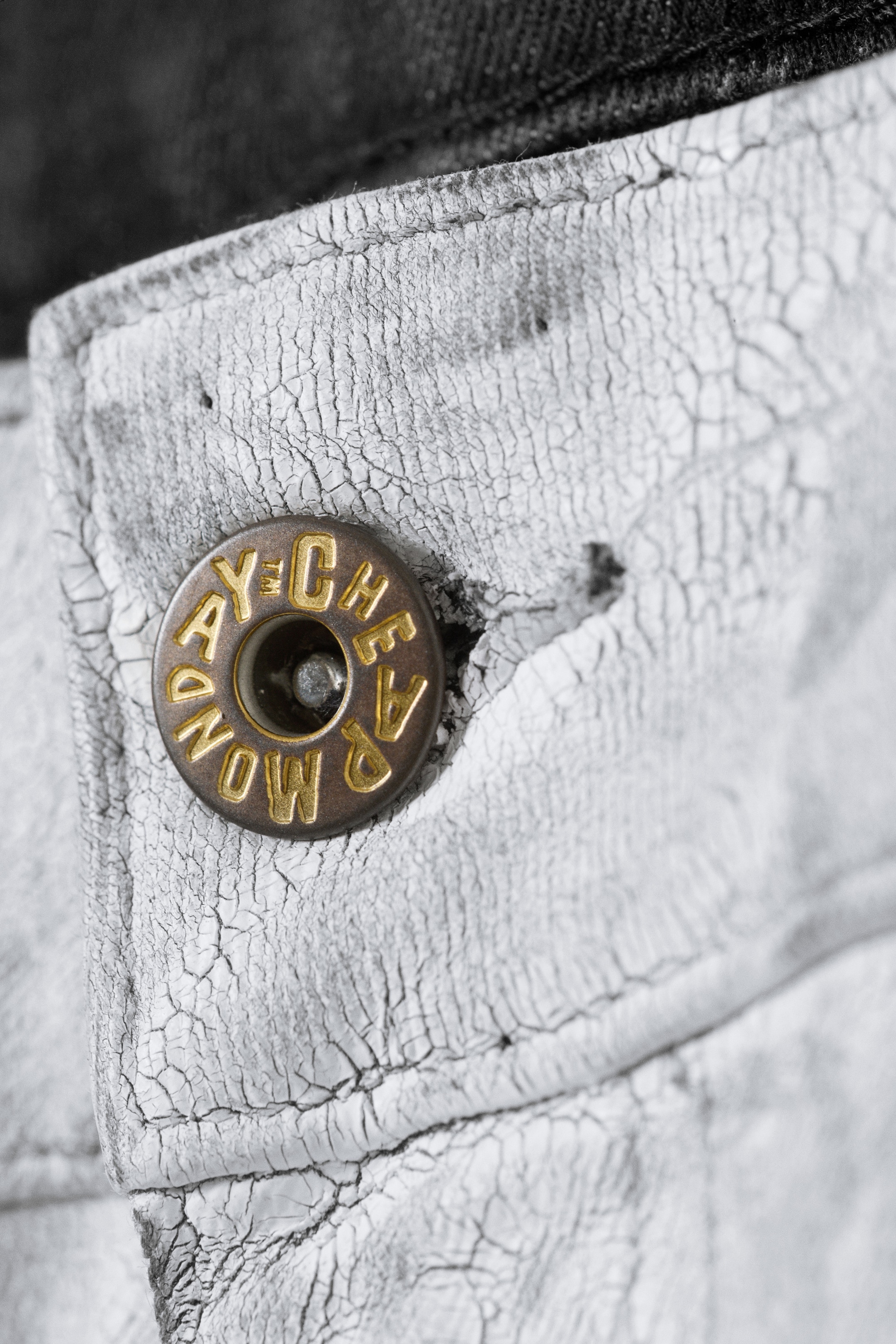 View larger image: A bronze-toned metal button, embossed with lettering, is fastened through a buttonhole on a piece of cracked off-white fabric. Visible stitching is detailing the fabric, and a dark grey fabric section is appearing along the top edge of the garment.