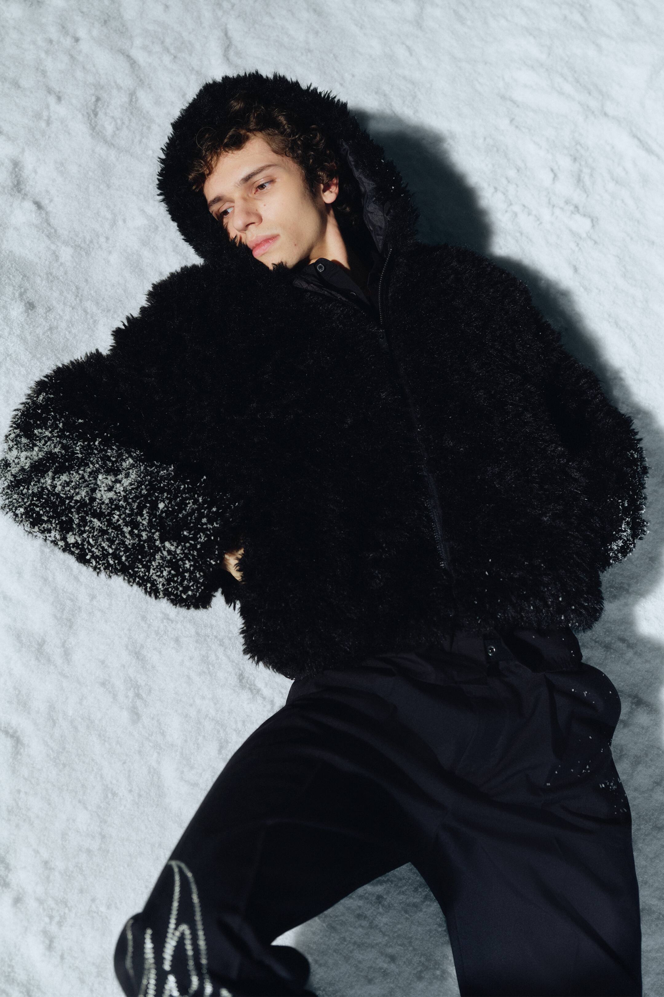 Ingrandisci l'immagine: Una giacca nera realizzata in materiale peloso presenta un cappuccio alzato e una chiusura con zip completa. Sono visibili pantaloni neri dal taglio ampio, che mostrano un motivo ondulato color argento sulla parte inferiore della gamba sinistra.