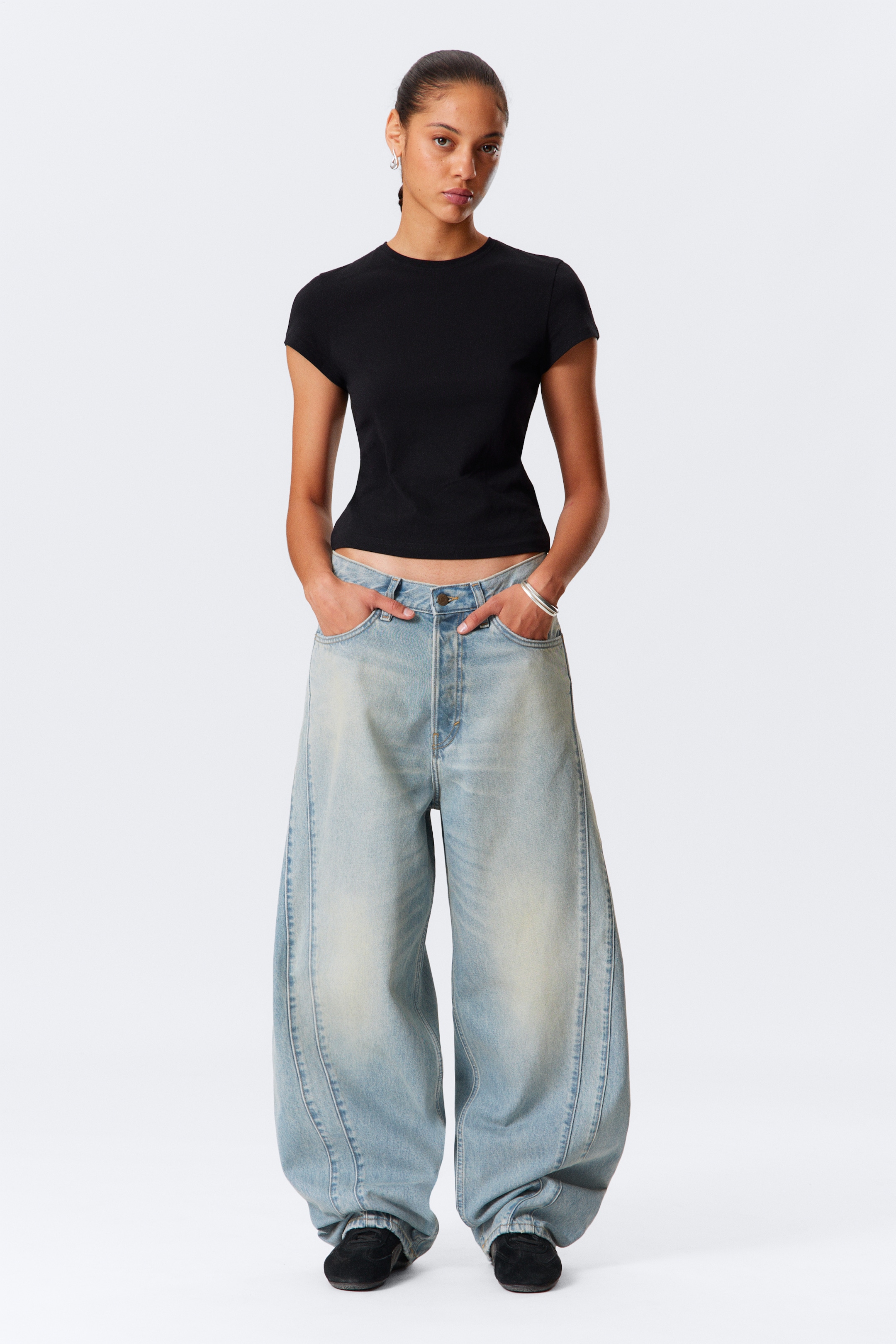 Visa större bild: En svart, figursydd, kortärmad topp med rund hals presenteras, dess fåll stannar ovanför midjan på ljusa, högmidjade, överdrivet vida jeans. Jeansen har framträdande vertikala sömmar längs framsidan och en blekt finish, vilket skapar en pösig silhuett.