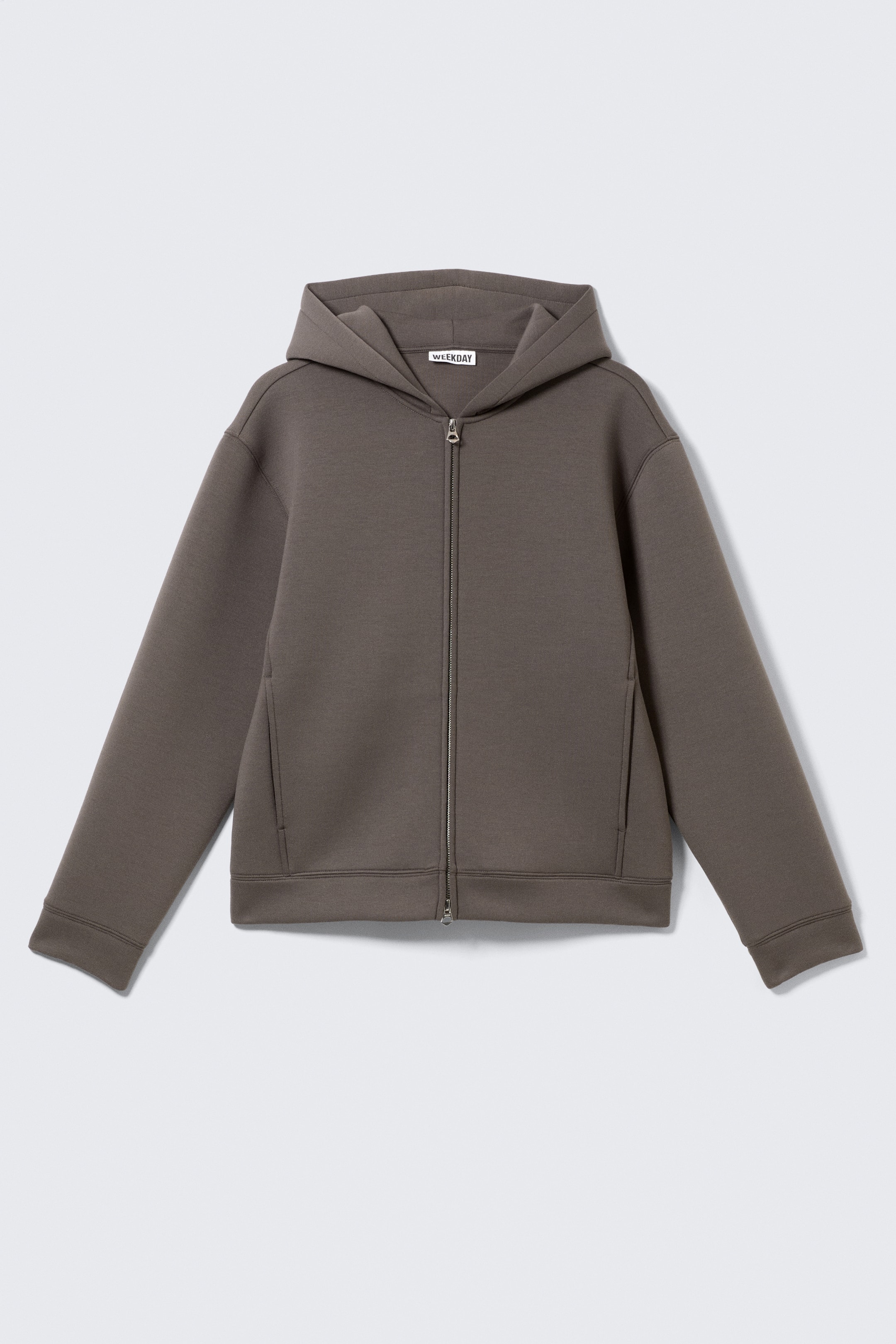 Grösseres Bild ansehen: Ein dunkelgrauer Kapuzenpullover mit durchgehendem Reißverschluss wird von vorne gezeigt, mit überschnittenen Schultern, einer strukturierten Kapuze und einem silberfarbenen Reißverschluss, der in der Mitte verläuft. Er ist mit zwei dezenten vertikalen Taschen an den Seiten gestaltet und mit schlichten Bündchen sowie einem sauberen Saum versehen.