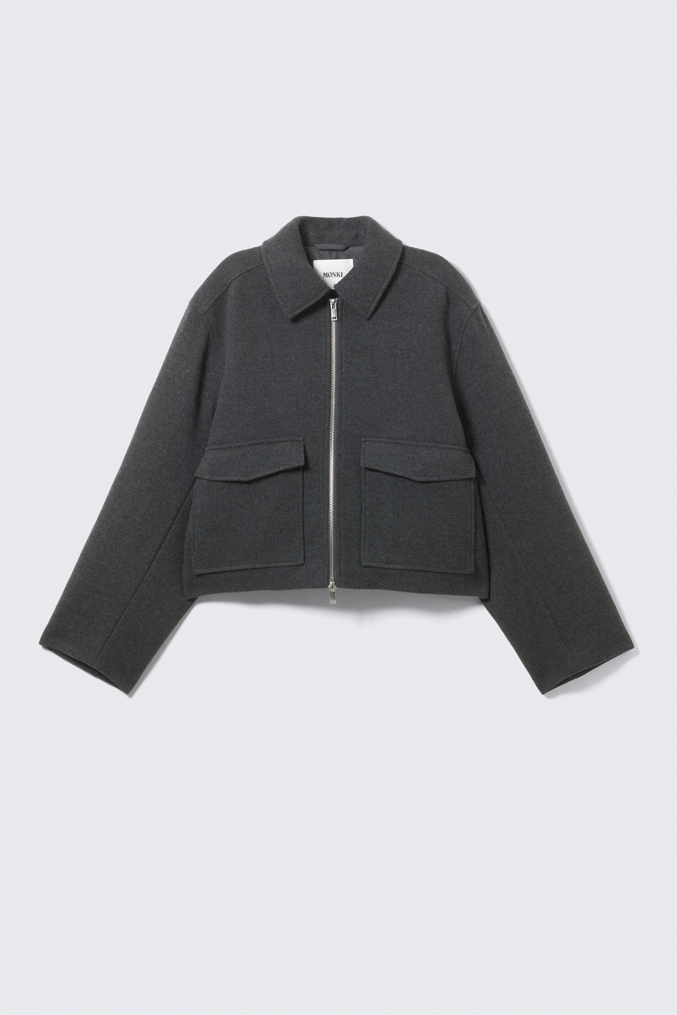Agrandir l'image: Une veste structurée et raccourcie est présentée à l'AVANT, avec une silhouette carrée dans un tissu texturé gris foncé. Elle est conçue avec un col plat et net, une fermeture éclair en métal à double sens de couleur argentée courant le long du centre avant, et deux grandes poches plaquées rectangulaires avec des rabats sur le bas avant. Les manches larges et droites se terminent par des poignets simples.