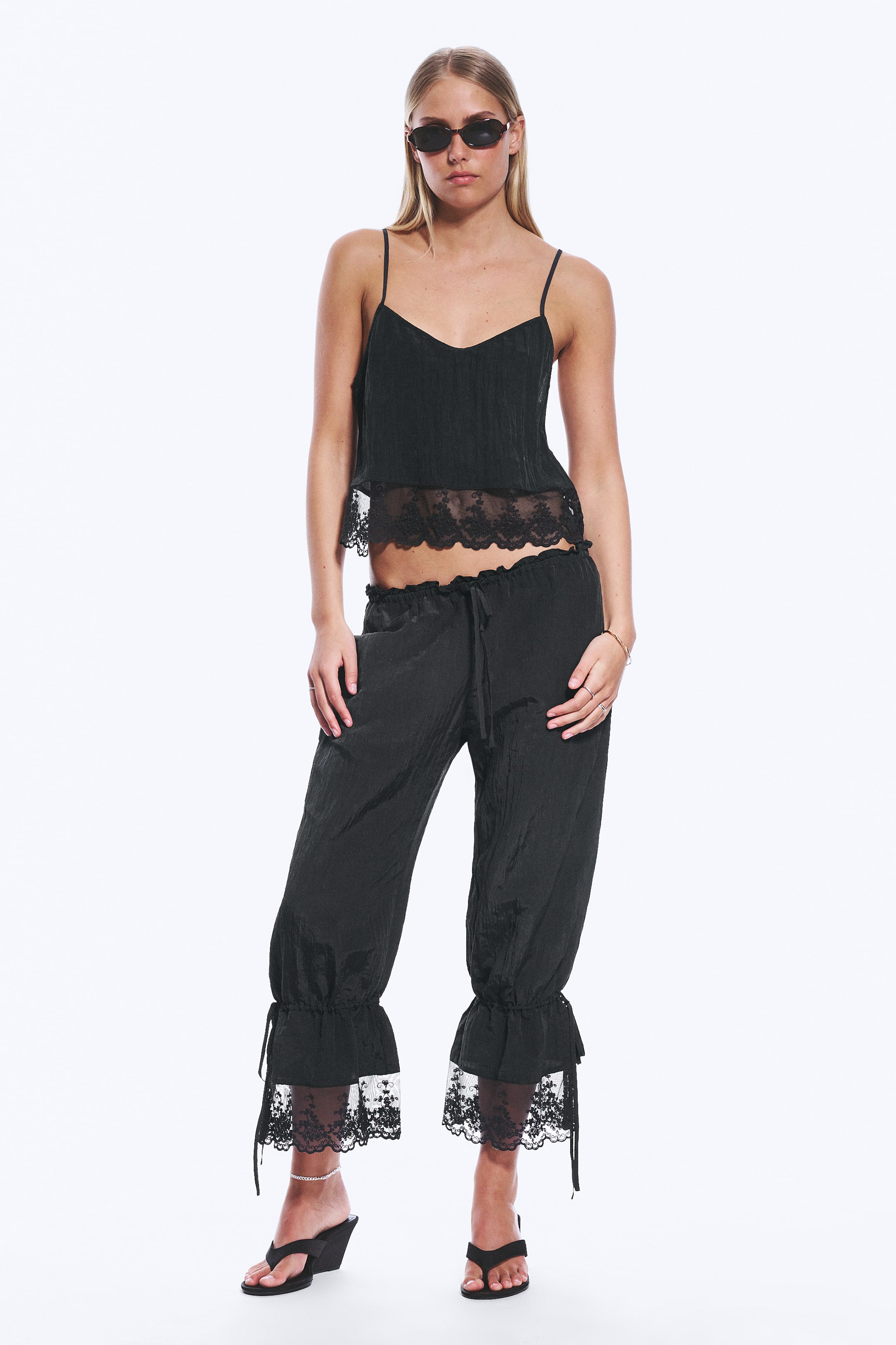 View larger image: The model is wearing black bloomers, characterised by a relaxed fit, a low-rise elasticated drawstring waistband, and side pockets where her hands rest. The three-quarter length legs conclude with wide, lace-trimmed hems, cinched with drawstrings tied into bows.