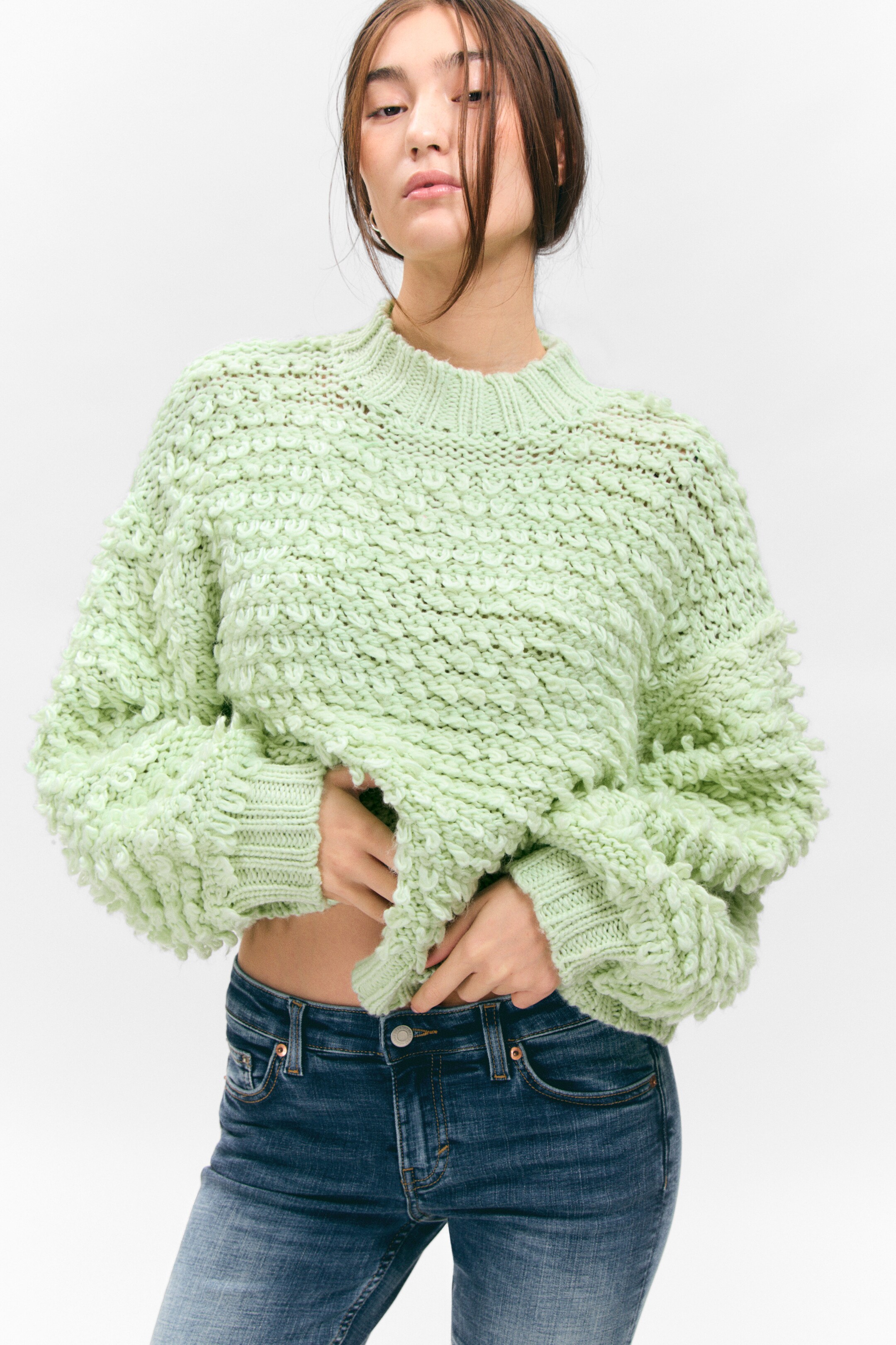 Se større billede: En lysegrøn chunky strikket sweater præsenteres forfra og har et tekstureret loopstrikmønster overalt. Den er designet med en høj ribbet mock-hals, lange voluminøse ærmer samlet i ribbede manchetter og en kort ribbet kant.
