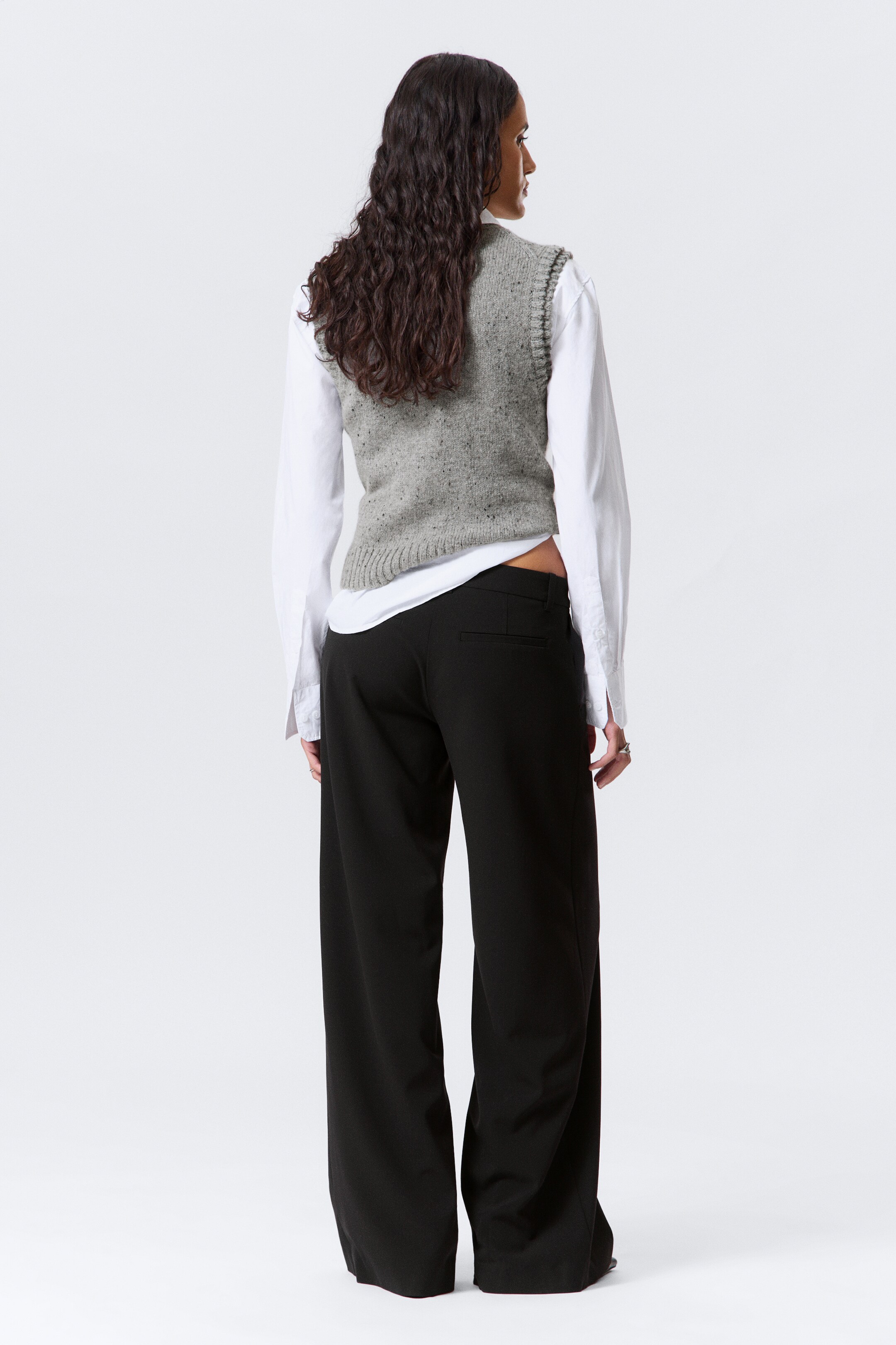 Visualizza immagine più grande: Un gilet a maglia grigio screziato, caratterizzato da giromanica e orlo a coste, è sovrapposto a una camicia bianca a maniche lunghe, con orlo e polsini che si estendono oltre il gilet. L'outfit è completato da pantaloni neri a gamba larga, che includono una visibile tasca posteriore, il tutto visto da dietro.