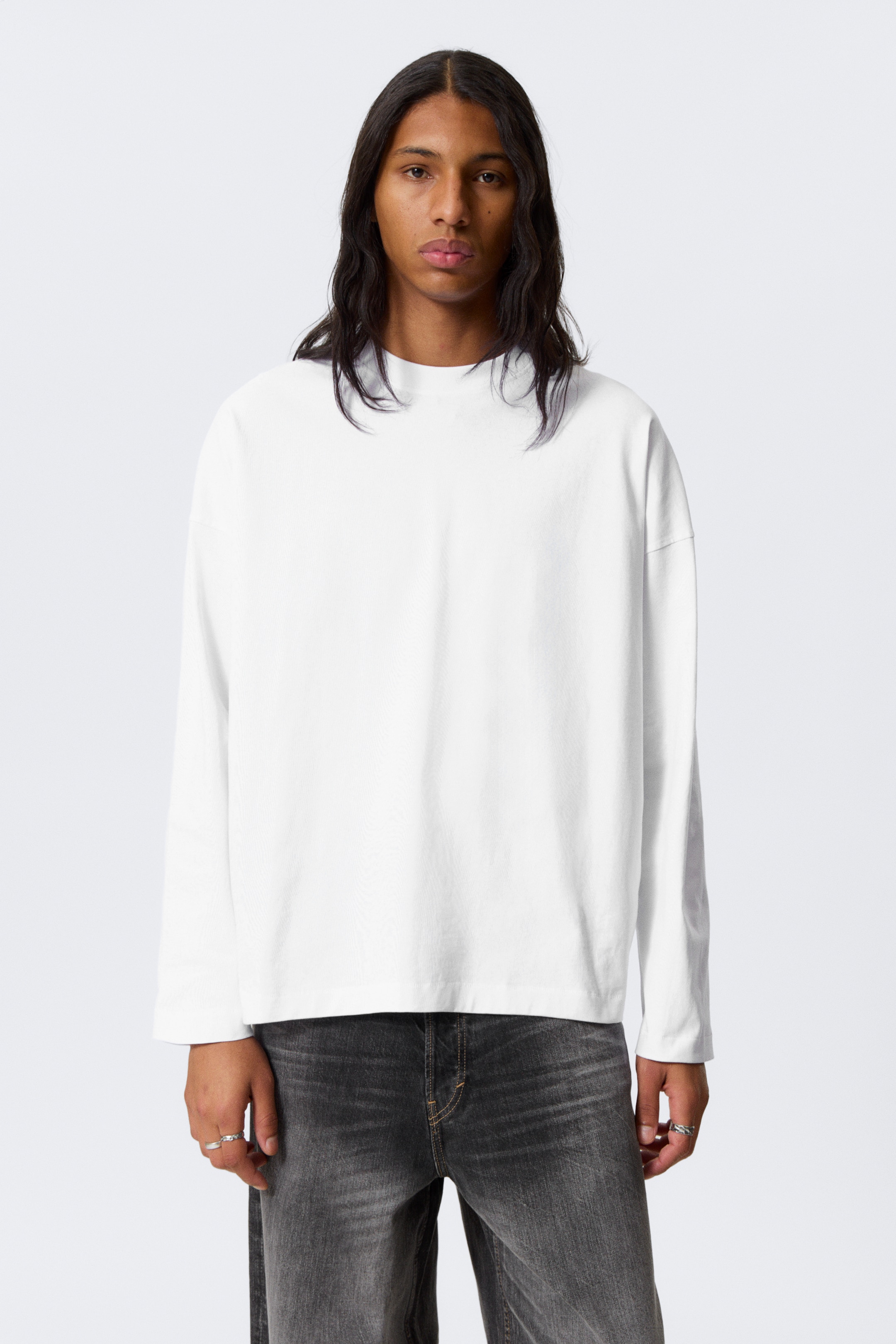 Visa större bild: En vit långärmad t-shirt visar en oversized passform, med nedhasade axlar och en rund hals. Dess raka fåll är synlig och ligger lager på lager över mörkt tvättade jeans.