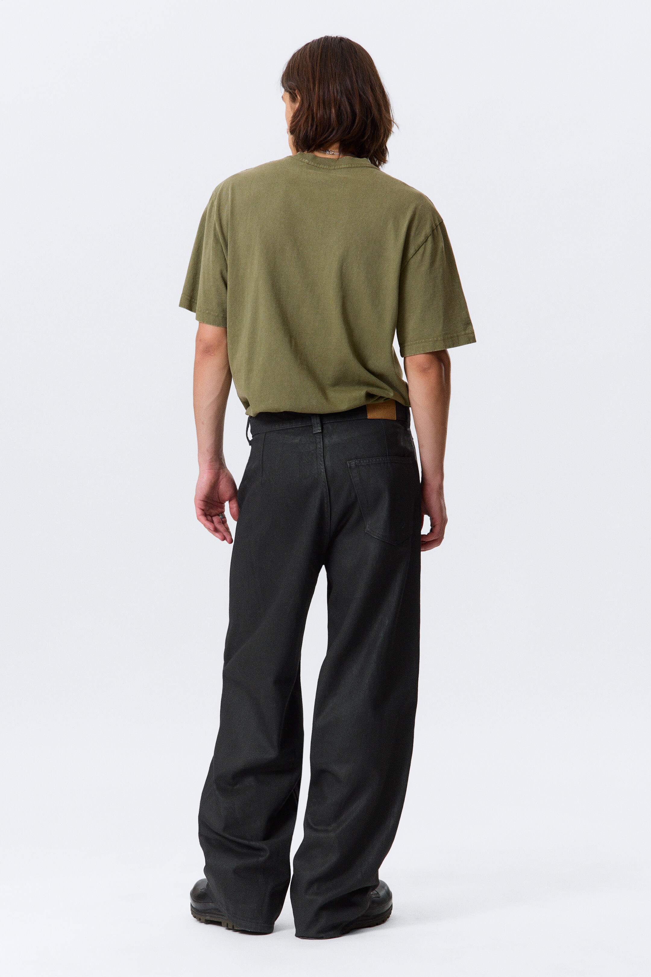 View larger image: Dark-toned, wide-leg pants are being worn from the back, featuring a high-rise waist, a subtle textured finish, and a light brown leather patch on the back right waistband. An olive green oversized t-shirt is being tucked into the pants.