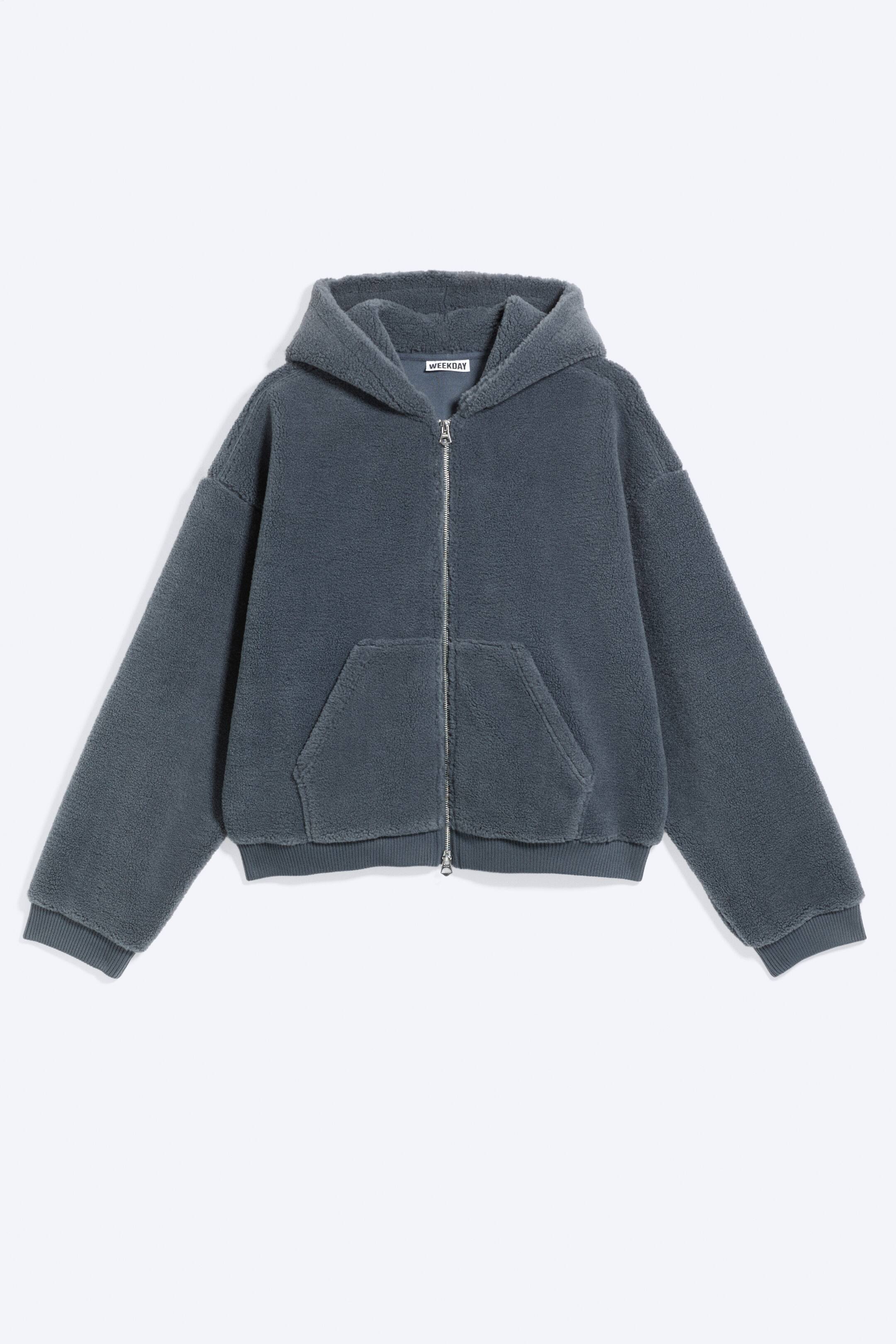 View larger image: A dark grey teddy fleece full-zip hoodie is shown from the front, featuring a relaxed, cropped fit. A hood sits at the top of the garment, and a silver-tone zip runs down the centre. Two front patch pockets are visible below, while ribbed cuffs and a ribbed hem complete the design, and a "WEEKDAY" tag is visible inside the back neck.