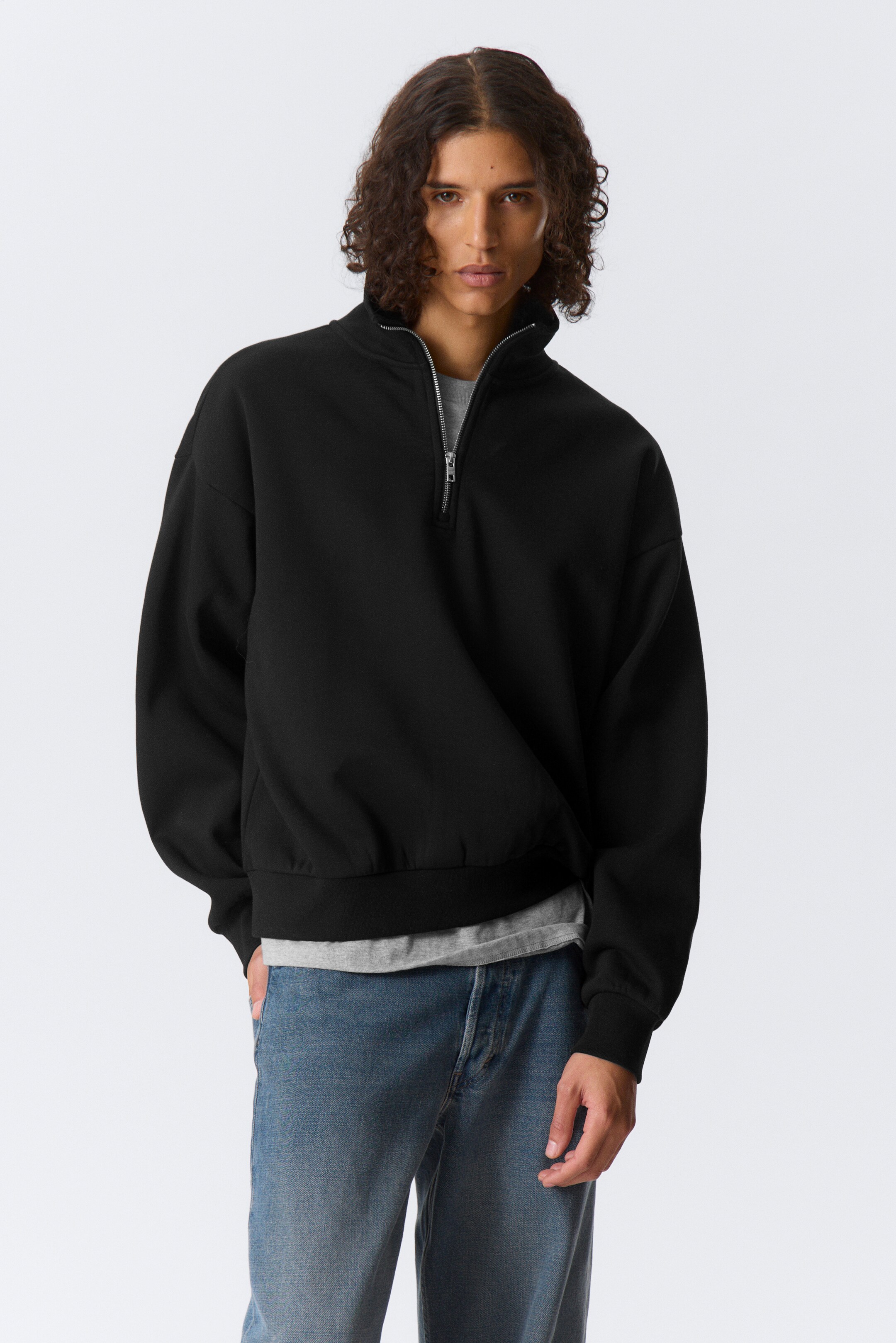 Ver imagen más grande: Un jersey negro con cremallera de un cuarto está mirando hacia adelante, con un cuello alto con una cremallera de tono plateado parcialmente desabrochada. Está mostrando un ajuste relajado con hombros caídos, puños acanalados y un dobladillo acanalado; se lleva una camiseta gris claro debajo.