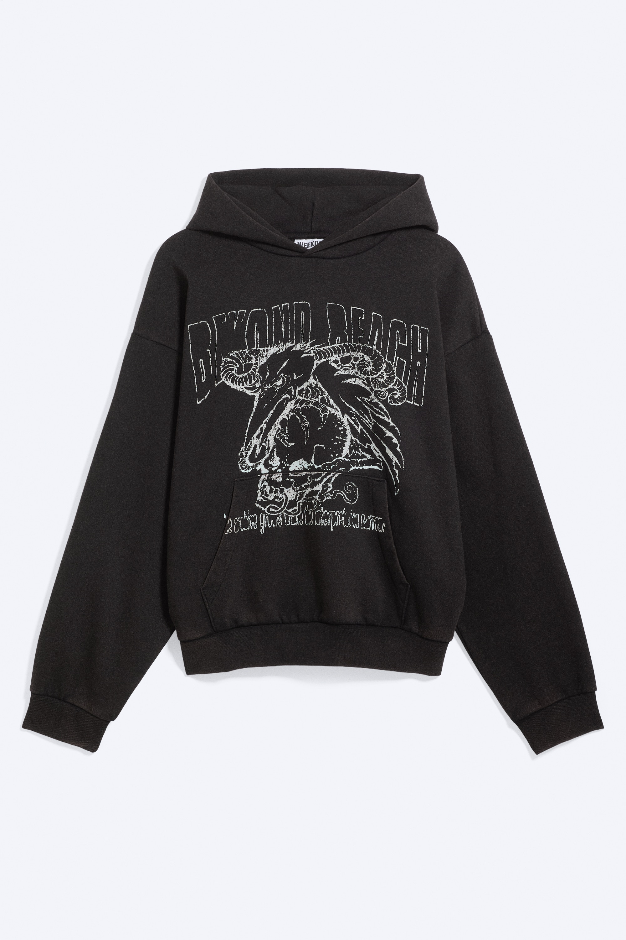 Een zwarte hoodie met losse pasvorm, voorzien van een grote witte grafische print over de borst, verlaagde schoudernaden en een ruime capuchon, wordt gedragen door het model. De hoodie is gelaagd over een grijs shirt dat zichtbaar is aan de zoom en kraag, en gecombineerd met olijfgroene ribfluwelen broek.