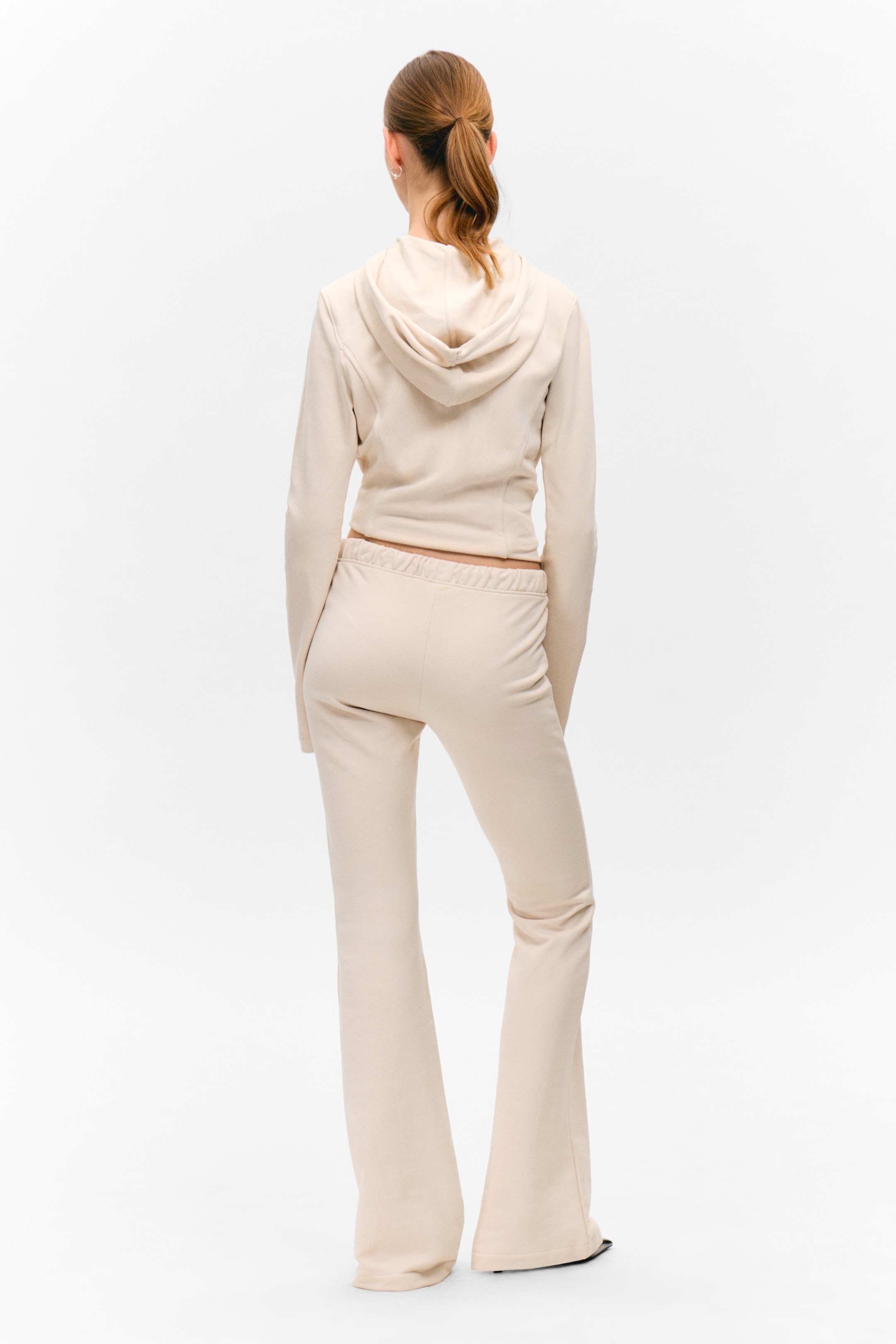 Vis større bilde: Et lyst beige joggedress vises fra baksiden, med en kort hettegenser og matchende utsvingte bukser. Hettegenseren har en hette og lange ermer, mens buksene har en elastisk midje og svinger betydelig ut fra kneet og ned.