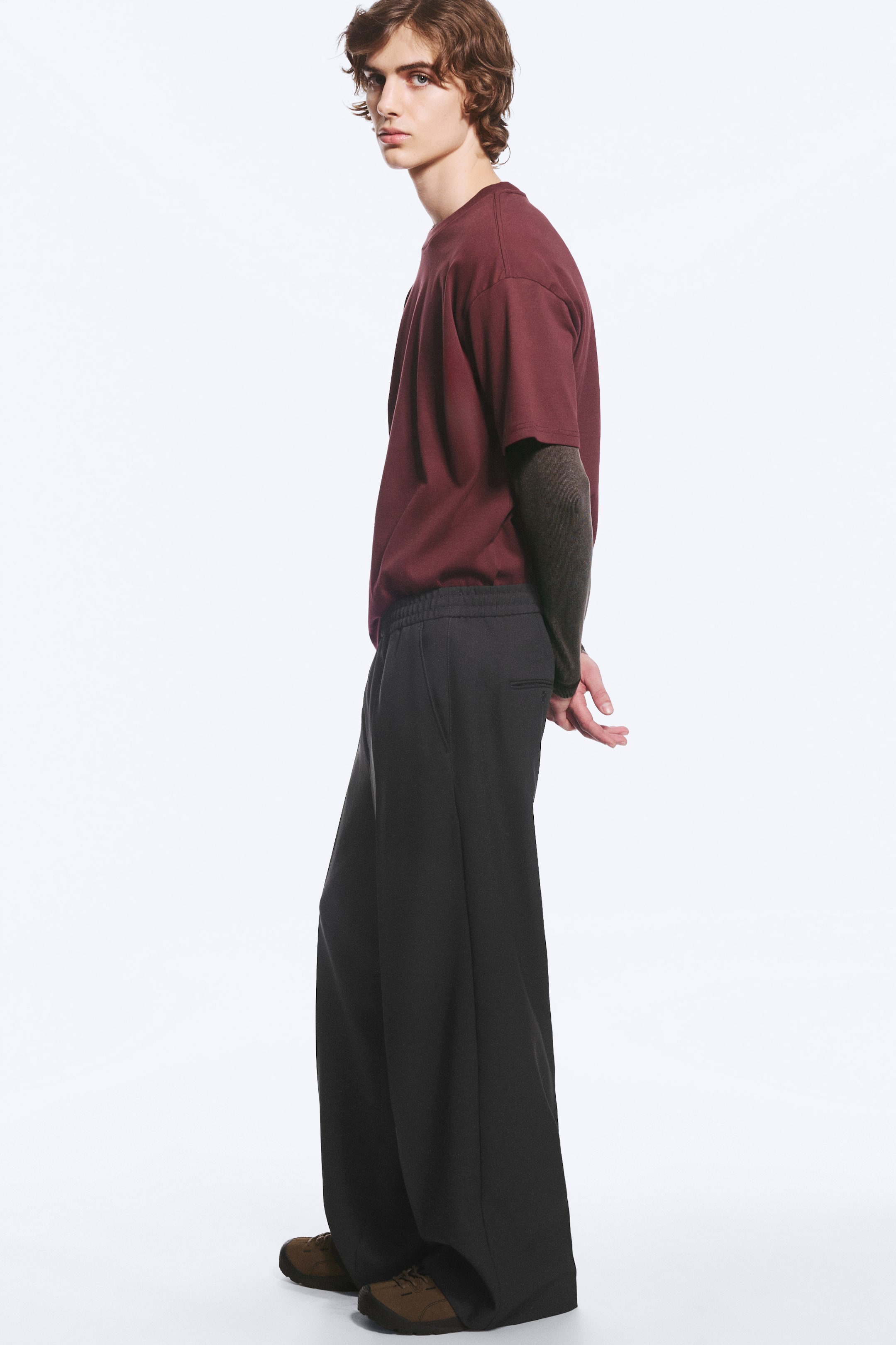Grotere afbeelding bekijken: De model draagt een losvallende, baggy pantalon in donkergrijs, met een wijde pijp, een gerimpelde tailleband en een paspelzak achter. De pantalon is gestyled met een gelaagd bordeauxrood T-shirt en bruine schoenen.