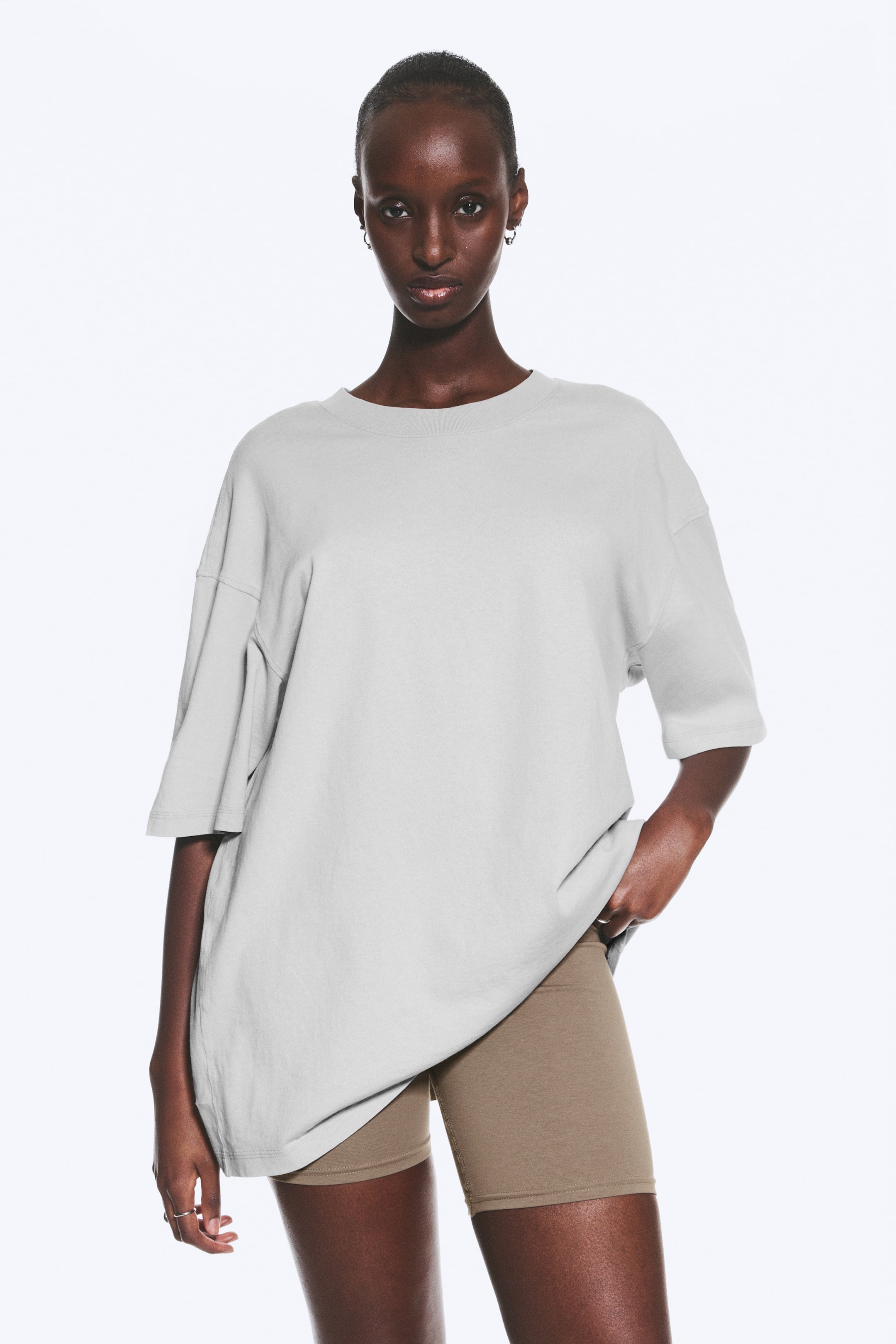 Se større billede: En lysegrå oversized t-shirt bæres fremadvendt. Den har en rund halsudskæring, sænkede skuldre og brede korte ærmer og falder løst ned over hofterne over brune cykelshorts.