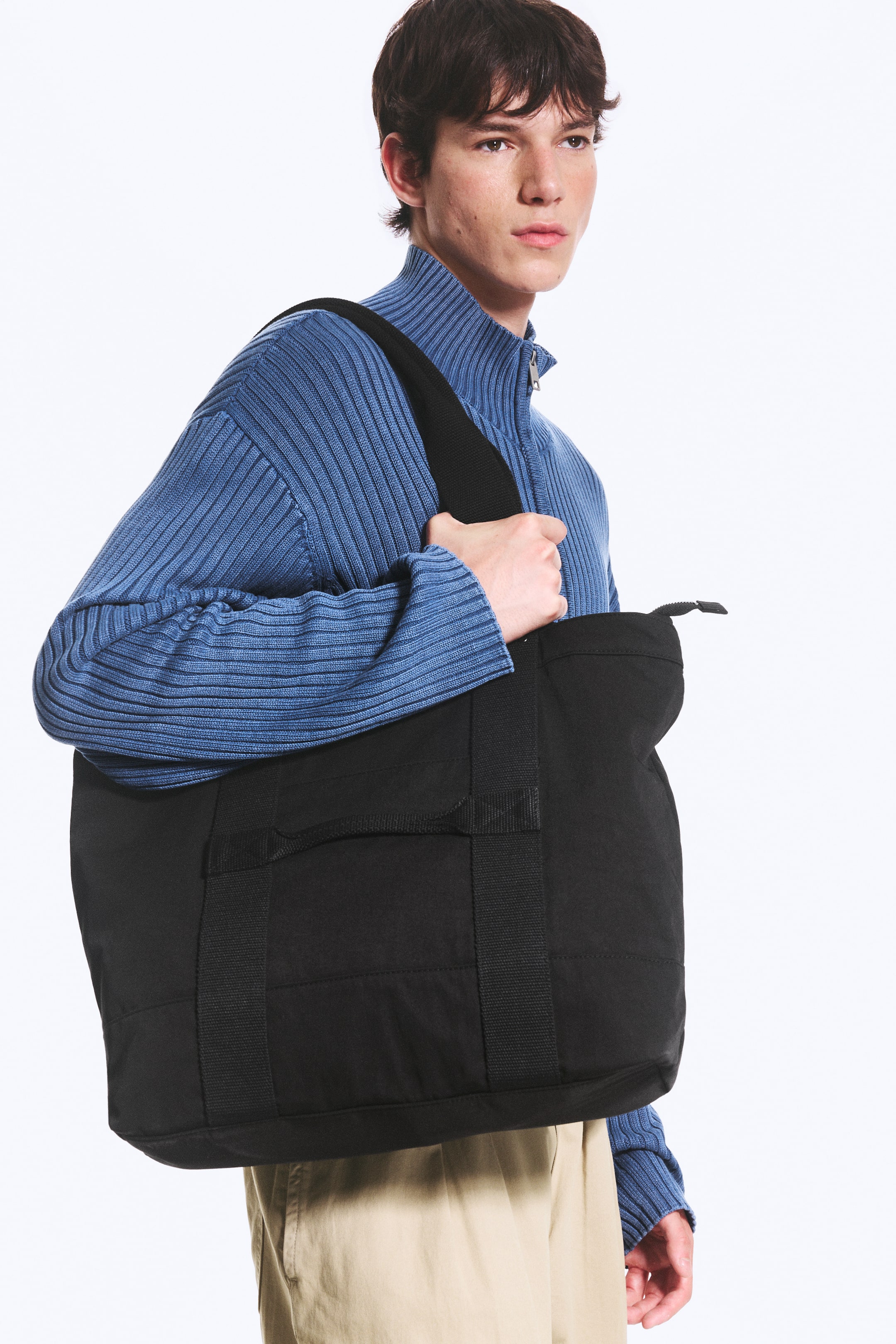Vezi imaginea mai mare: O geantă tote neagră cu un design matlasat, buzunare externe și închidere cu fermoar în partea de sus este purtată de model. Modelul ține cureaua lată, care se sprijină peste un pulover albastru cu nervuri și fermoar pe jumătate.