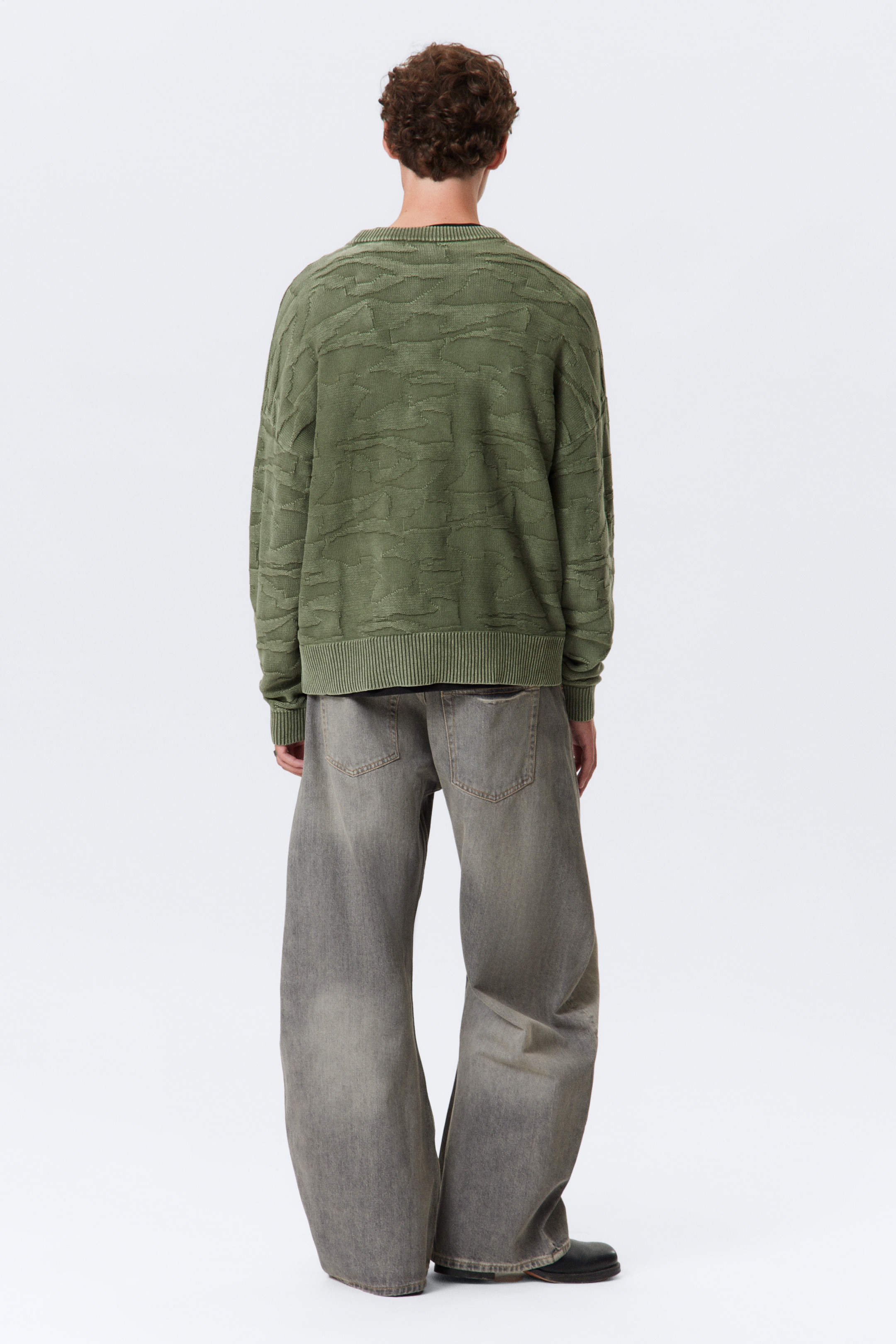 Agrandir l'image: Un pull vert olive, avec un motif texturé abstrait et des ourlets et poignets côtelés, est présenté de dos. Il est porté avec un jean en denim gris délavé à jambes larges, les deux vêtements affichant une coupe décontractée.