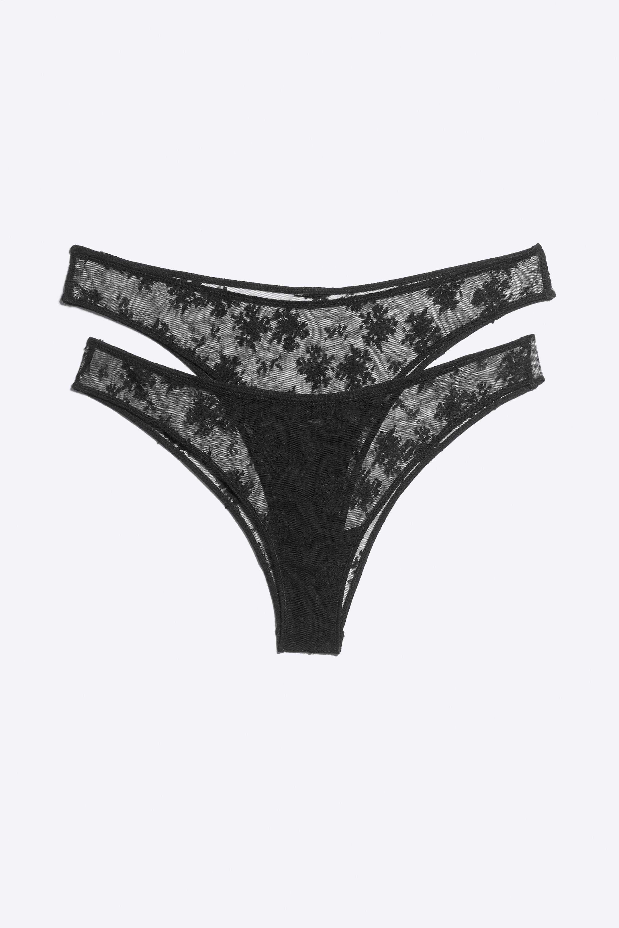 Größeres Bild ansehen: Zwei schwarze Tanga-Slips sind in der Vorderansicht dargestellt und verfügen über ein transparentes Mesh-Gewebe mit aufwendiger floraler Spitzenstickerei über die gesamte Oberfläche. Jeder Tanga ist mit einem hoch geschnittenen Beinausschnitt und einem doppelt gerafften Bund gestaltet, wodurch an den Hüften ein Cut-out-Effekt entsteht.
