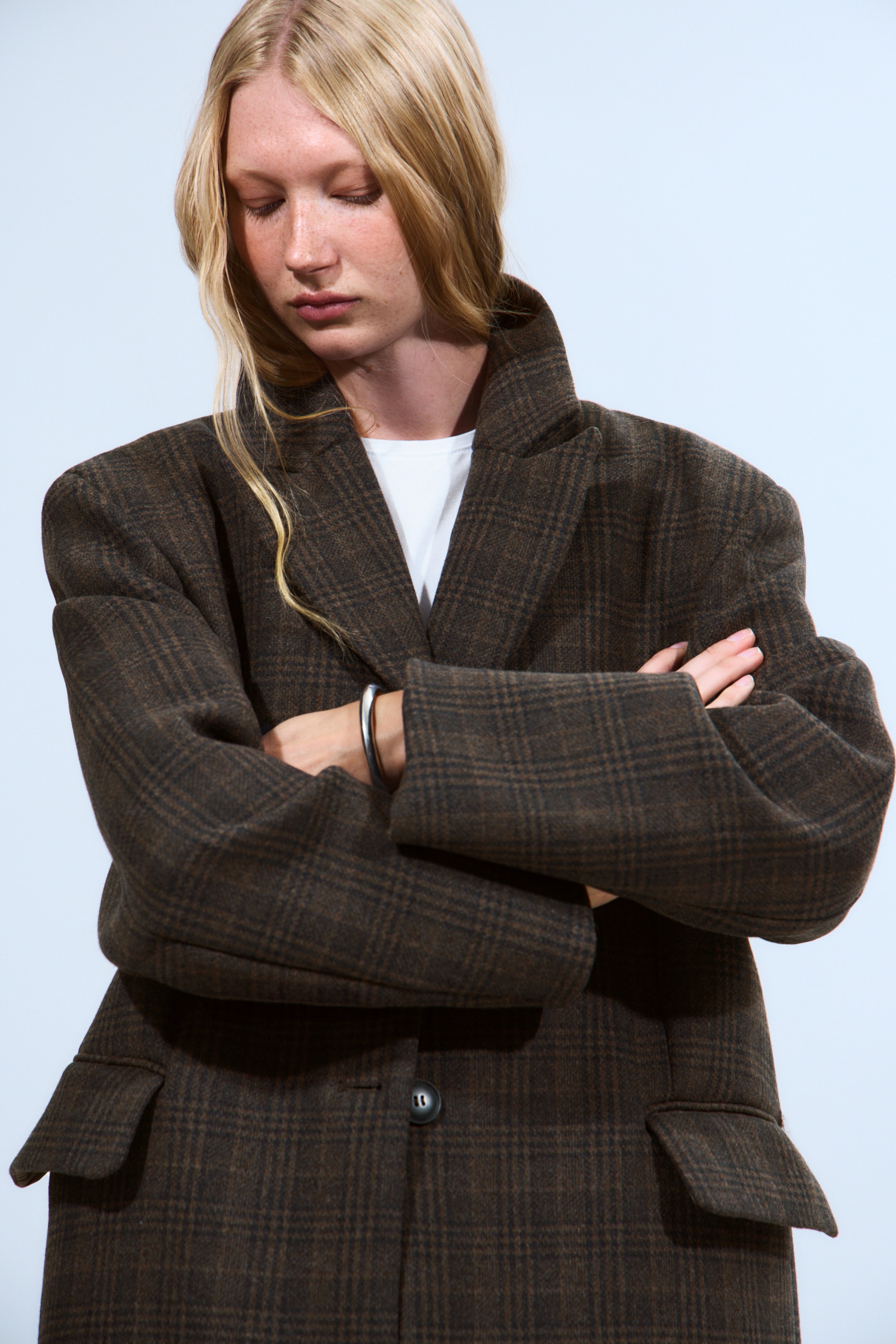 Größeres Bild ansehen: Ein brauner karierter Blazer wird von vorne präsentiert, leicht nach rechts geneigt, um seine strukturierte, übergroße Passform mit breiten Schultern und breiten Kerbrevers hervorzuheben. Er zeigt auch einen einzelnen dunklen Knopfverschluss und zwei große Pattentaschen an der unteren Vorderseite und wird über einem weißen Rundhalshemd getragen.