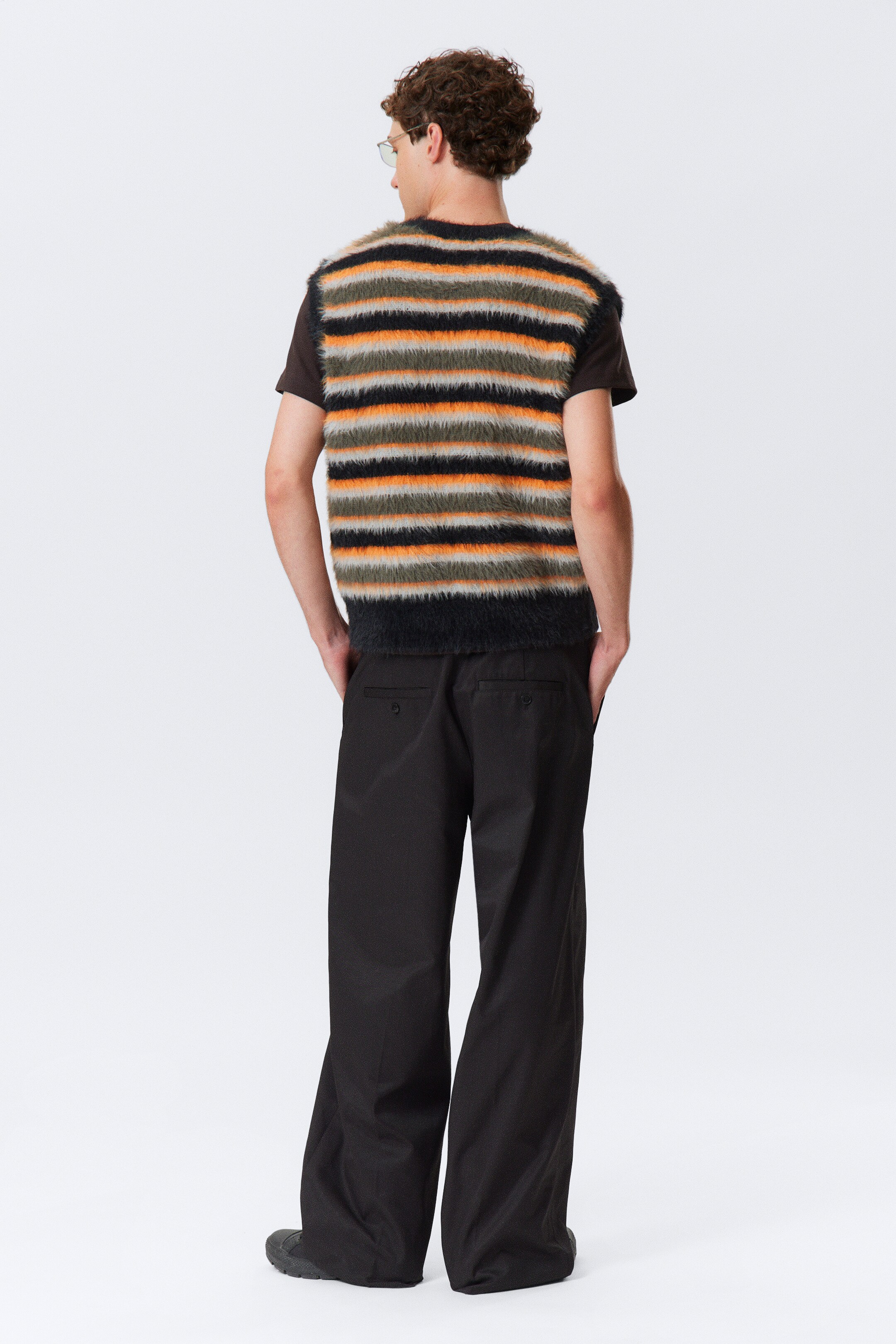 Agrandir l'image: Un gilet duveteux à rayures horizontales, avec des rayures noires, orange, vert olive et gris clair, est porté par-dessus un t-shirt à manches courtes foncé. Le gilet est assorti à un pantalon ample à jambes larges de couleur foncée, visible de dos, montrant deux poches arrière.