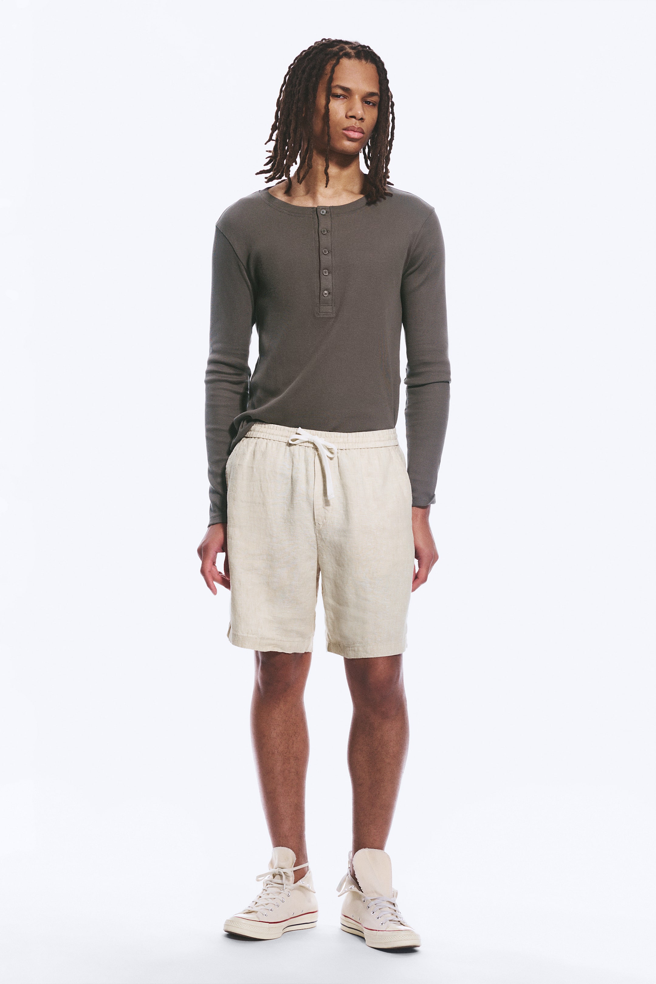 Ver imagen más grande: Una camiseta henley de manga larga en gris oscuro presenta una tapeta de cinco botones y un escote redondo. Se lleva metida en unos pantalones cortos de lino beige claro con cordón, que muestran una cintura elástica con un cordón blanco roto atado y bolsillos laterales.