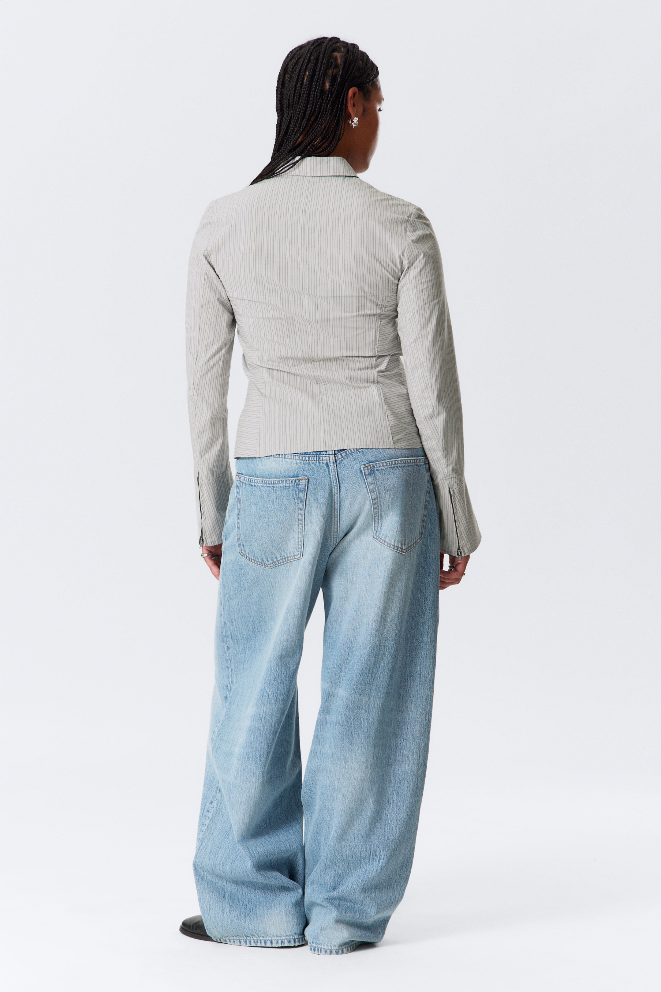 Grösseres Bild ansehen: Ein hellgraues, gestreiftes Oberteil und hell gewaschene, weit geschnittene Jeans werden getragen, von hinten betrachtet. Das figurbetonte Oberteil zeichnet sich durch einen spitzen Kragen, taillierte Nähte, die die Taille betonen, und lange Ärmel mit ausgestellten, geteilten Manschetten aus. Die locker sitzenden Jeans sammeln sich um die Knöchel.