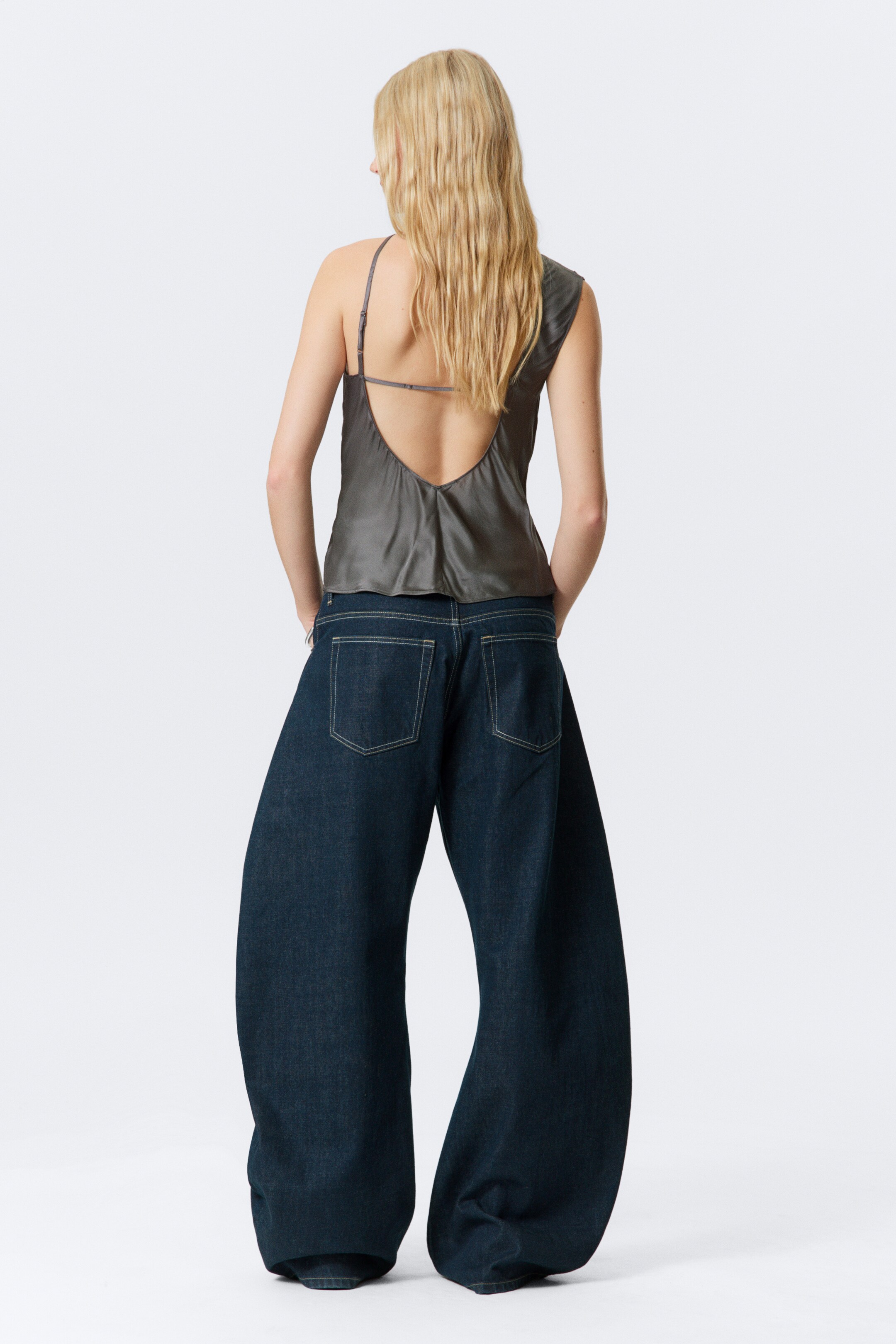 View larger image: A dark gray satin-like top and dark wash jeans are being shown from the back, highlighting the top's low U-shaped back with one thin adjustable strap and one wider strap. The wide-leg jeans are presenting a loose fit, detailed with two large back patch pockets and contrasting white stitching.