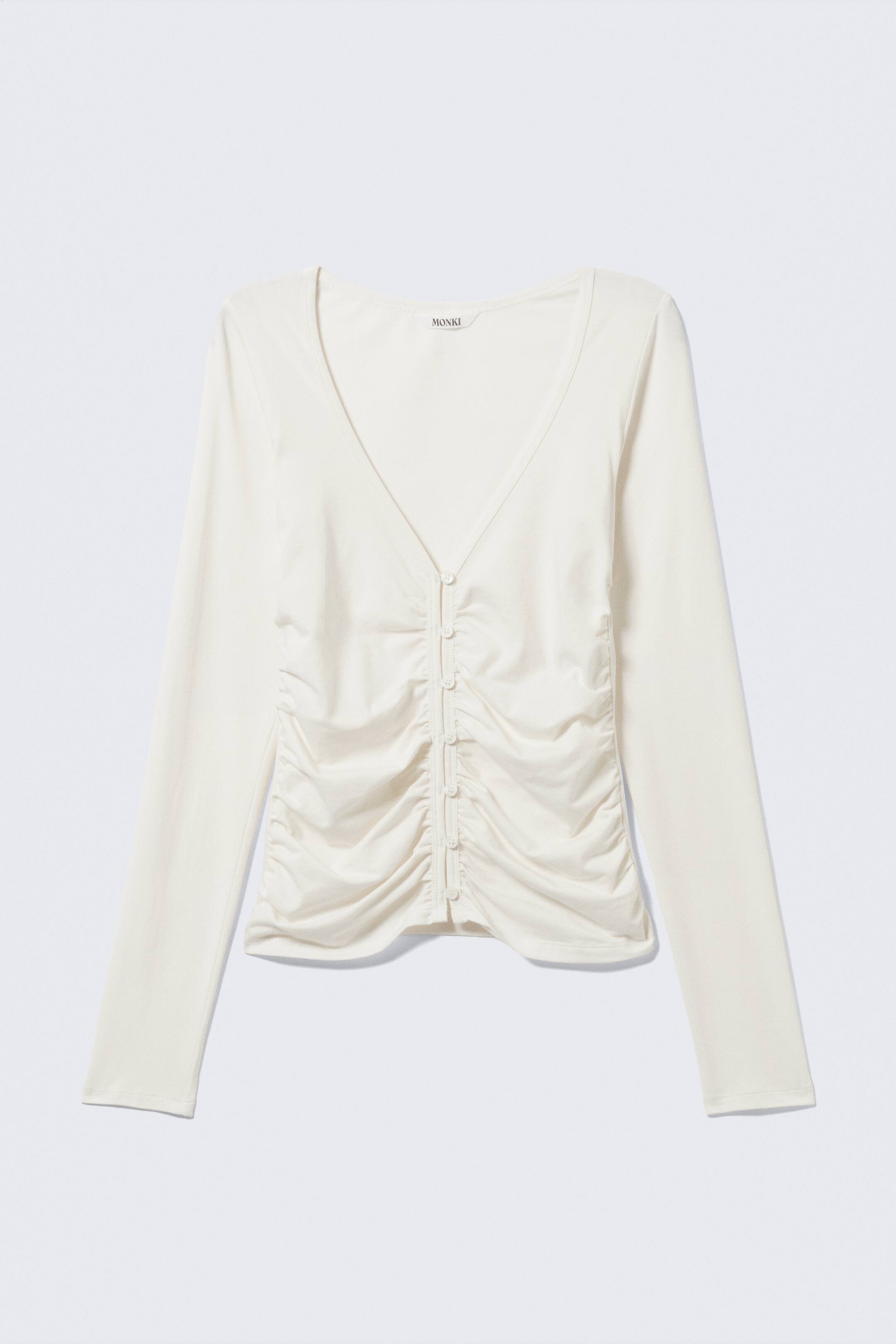 Zobacz większy obraz: Kremowy top jest skierowany PRZODEM, posiada dekolt w kształcie litery V oraz długie rękawy. Kolumna małych, okrągłych guzików biegnie w dół środka przodu, otoczona pionowym marszczeniem zbierającym tkaninę po obu stronach listwy.