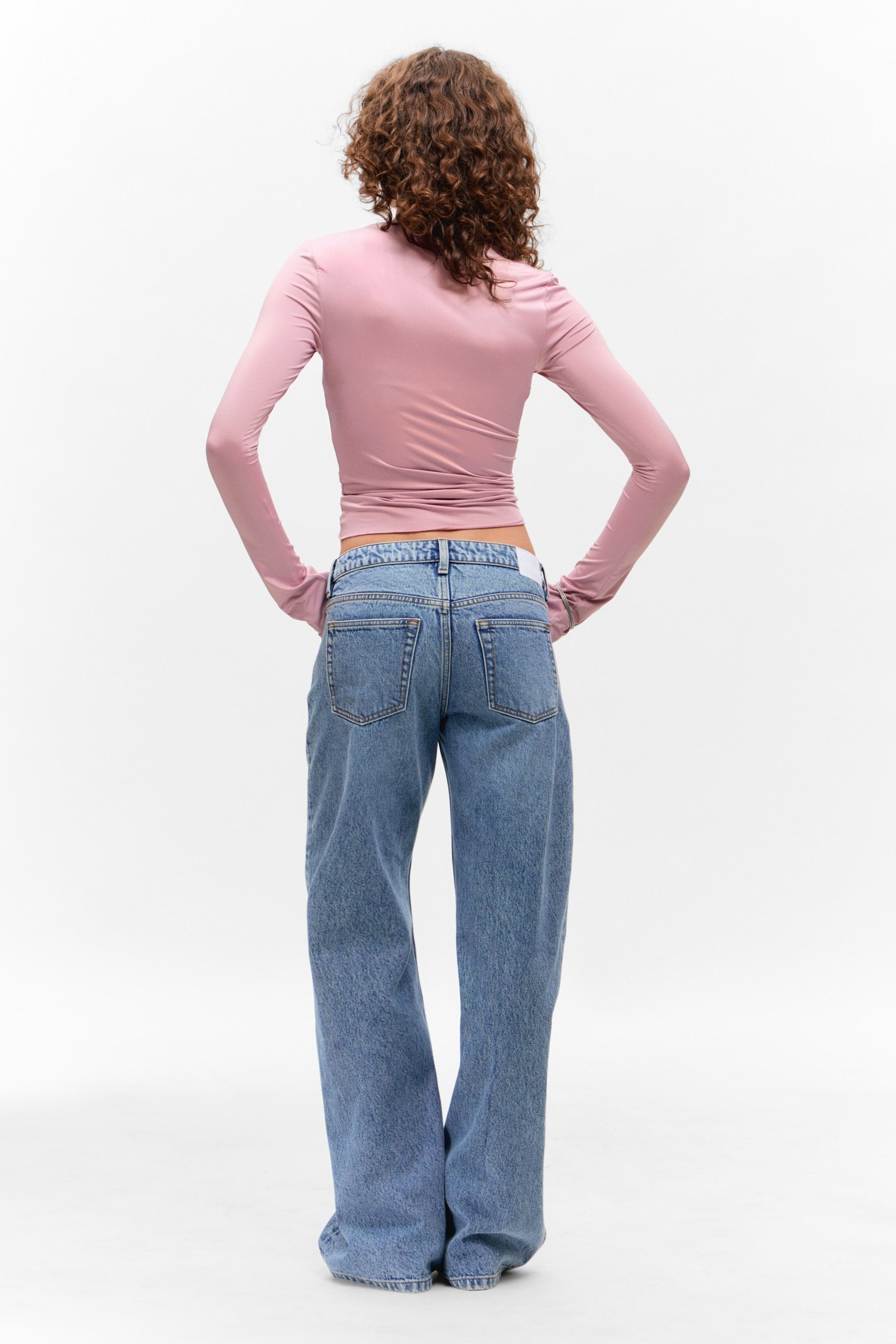 Größeres Bild ansehen: Ein hellrosa, langärmliges Oberteil mit hohem Kragen wird getragen, das mit subtilen Raffungen am Saum eng anliegend erscheint. Es wird mit hellblauen, weit geschnittenen Jeans kombiniert, die eine entspannte Passform und sichtbare Gesäßtaschen aufweisen. Die Produkte werden von hinten gezeigt.