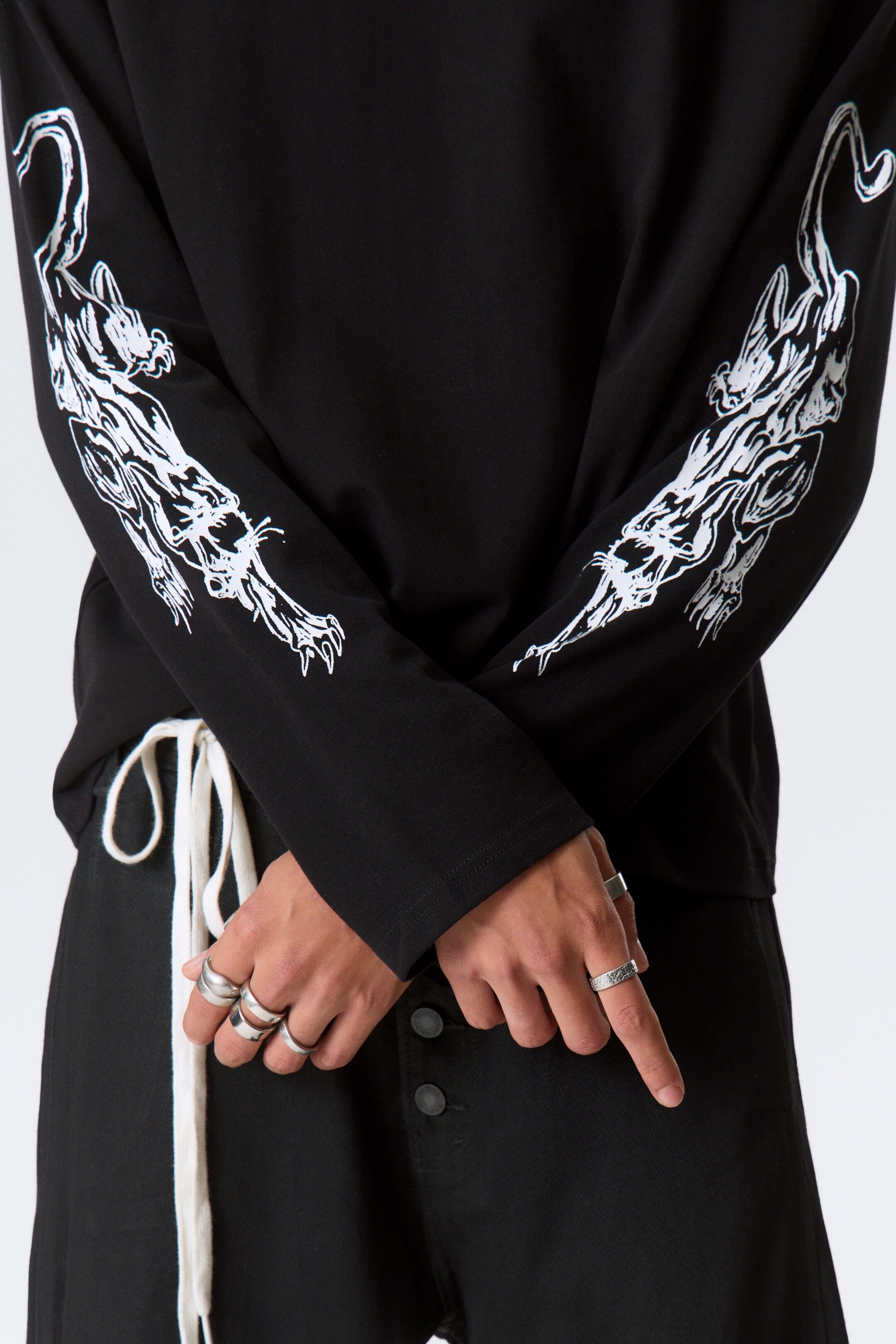 Vezi imaginea mai mare: Un tricou negru cu mânecă lungă afișează modele grafice albe cu feline stilizate întinse de-a lungul ambelor mâneci. Tricoul prezintă manșete simple, tivite și este văzut dintr-o perspectivă frontală, ușor înclinată.