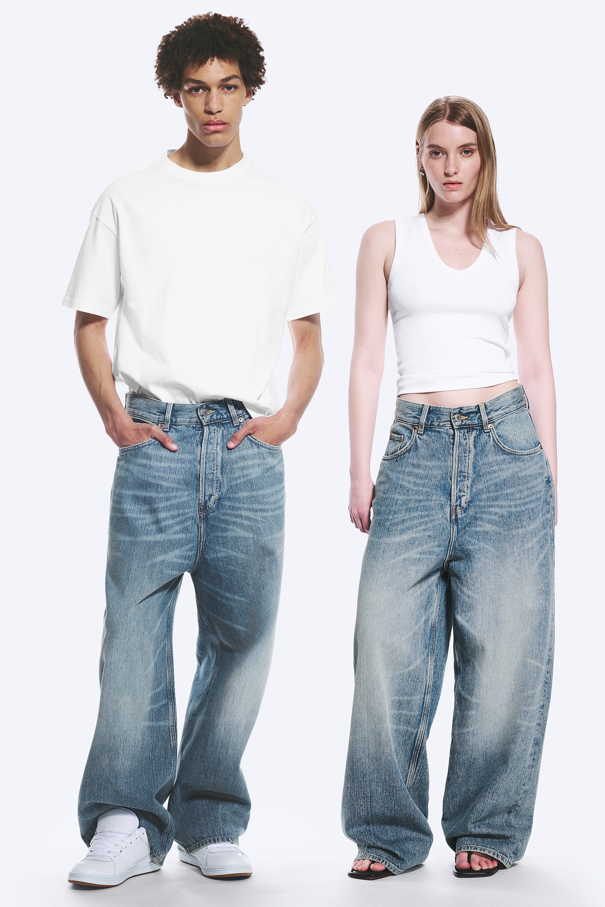 Un jean bleu clair à jambes larges est présenté de face, avec une coupe taille haute, une fermeture à boutons et deux poches avant en forme de cuillère avec une poche à monnaie. Le denim présente un délavage décoloré avec des moustaches sur les cuisses.