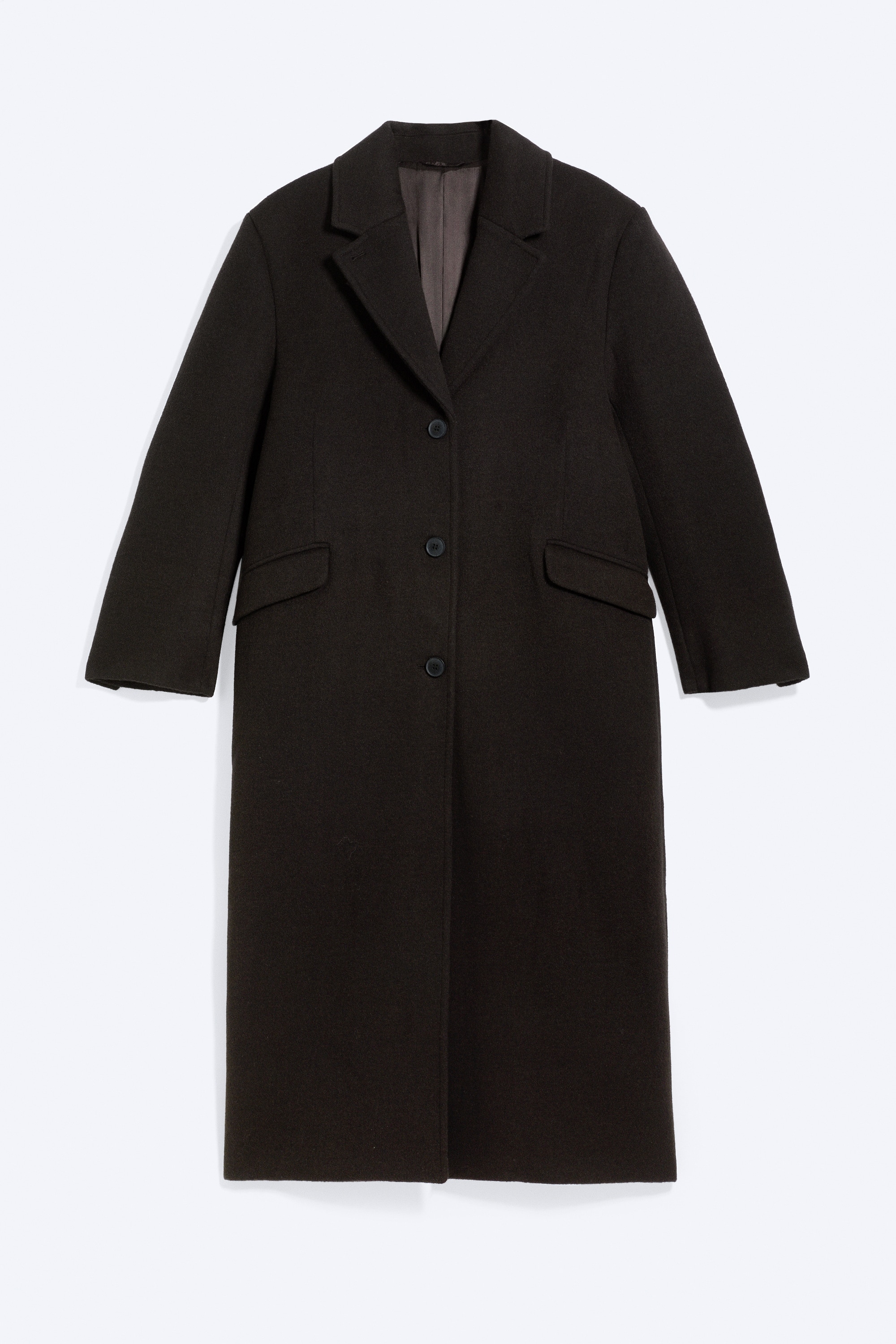 En oversized, lang mÃ¸rkebrun car coat bÃ¦res Ã¥bent af modellen og har markerede skuldre, revers med hak og knapdetaljer foran. Frakken er stylet over en mÃ¸rkeblÃ¥ hoodie med lynlÃ¥s og matchende sweatpants.