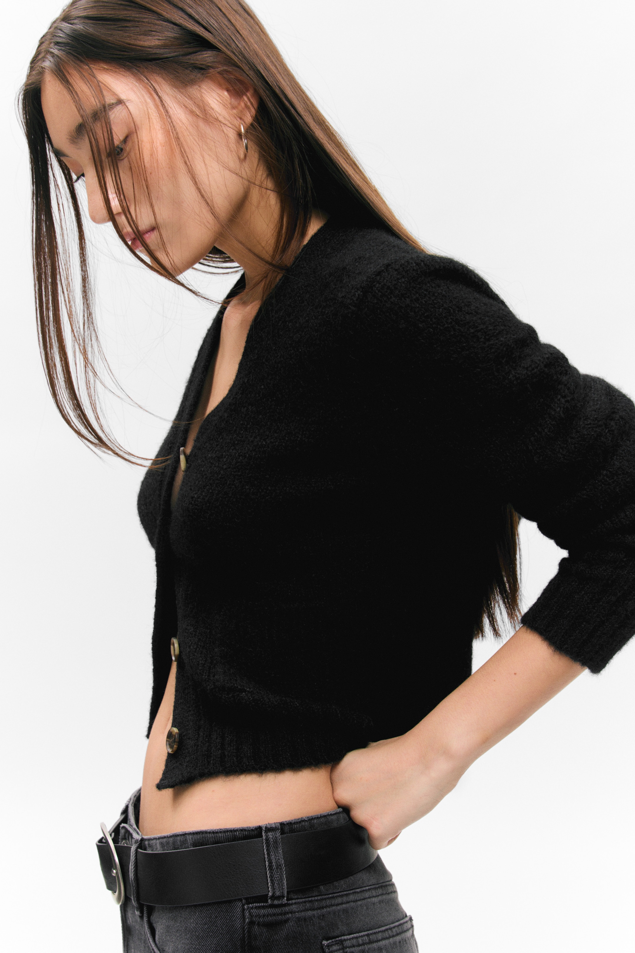 Größeres Bild ansehen: Eine schwarze V-Ausschnitt-Strickjacke mit flauschiger Textur und hellen Knöpfen an der Knopfleiste wird von dem Model getragen. Die kurz geschnittene Strickjacke endet oberhalb der Taille, und das Model steckt eine Hand in den Bund ihrer dunklen Hose in der Nähe des Saums der Strickjacke.