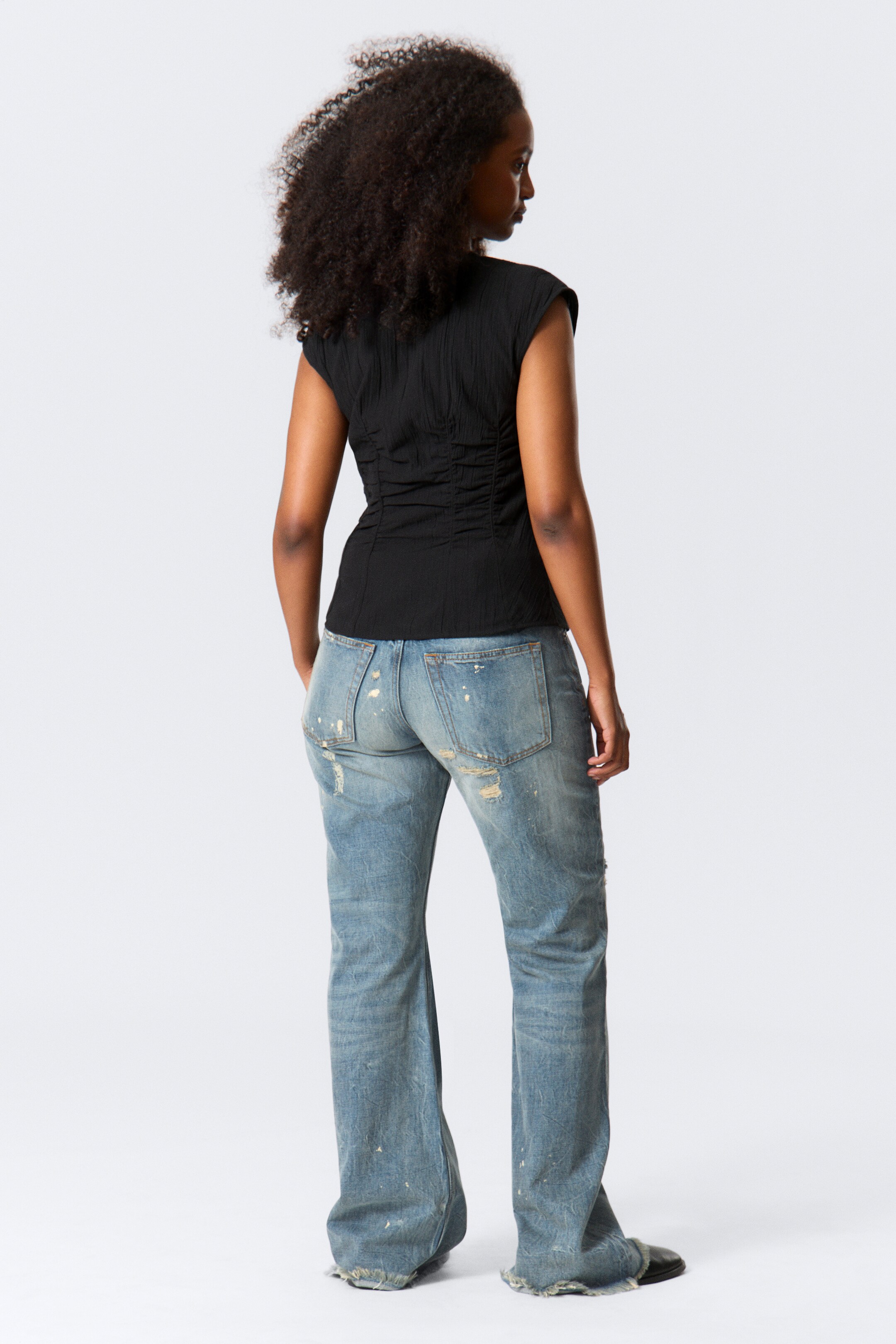 Grotere afbeelding bekijken: Een zwart getextureerd topje wordt van de achterkant getoond, met kapmouwtjes en verticale rimpelingen langs de achterpanelen, wat een aansluitend silhouet creëert. Het wordt gestyled met lichtblauwe, versleten bootcut jeans die scheuren, verfvlekken en rafelige zomen hebben.