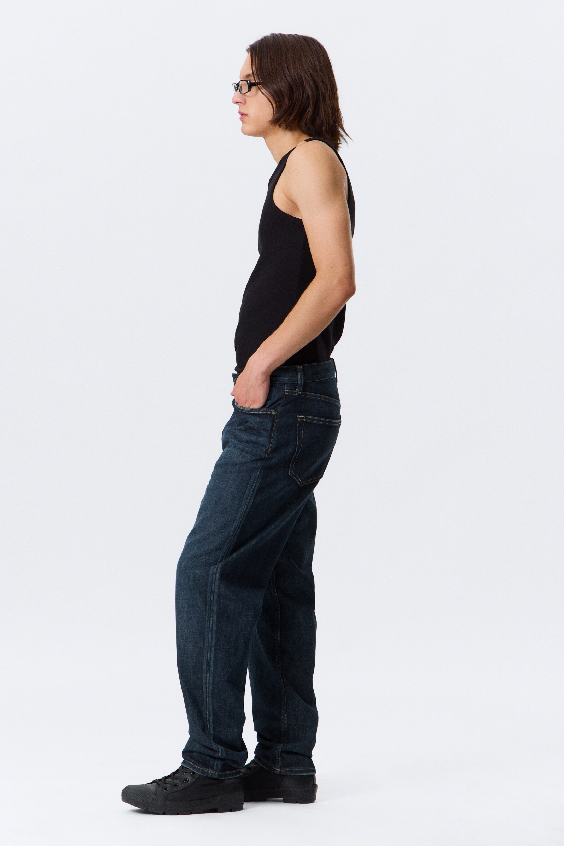 View larger image: Dark blue relaxed-fit jeans are being presented from a side profile, featuring prominent front leg seams, five-pocket styling, and a full length pooling at the ankles. A black fitted tank top is being worn tucked into the jeans.