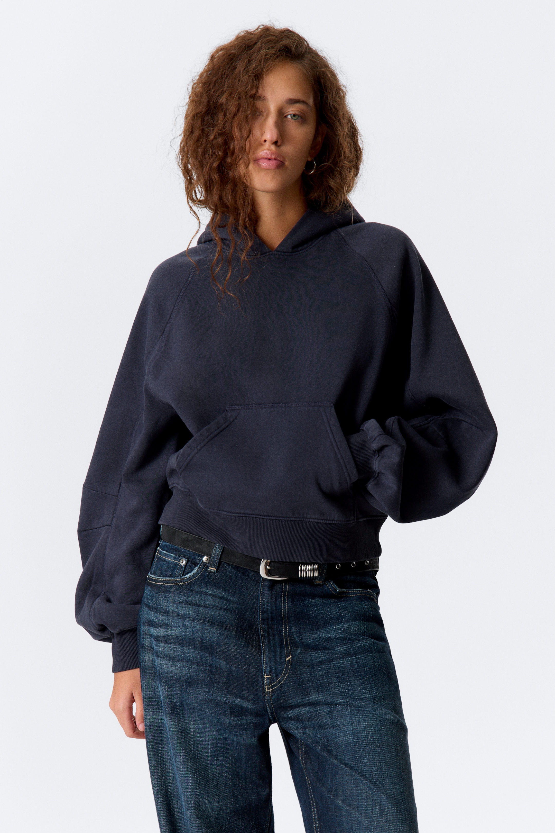 Größeres Bild ansehen: Ein gewaschener marineblauer Hoodie wird von vorne präsentiert und verfügt über eine gekürzte, übergroße Passform mit paneelförmigen Raglanärmeln, die voluminös sind und in gerippten Bündchen enden. Es zeigt eine große Kängurutasche auf der Vorderseite und einen gerippten Saum.