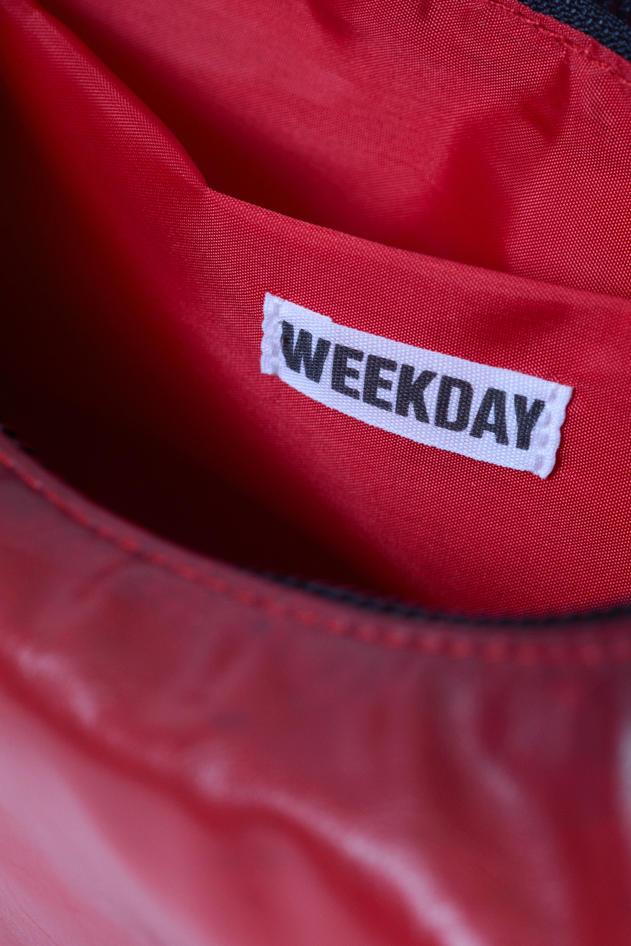 Agrandir l'image: L'intérieur d'un sac à main est montré, avec une doublure en tissu rouge. Une poche plaquée interne blanche avec 'WEEKDAY' imprimé en lettres majuscules noires est visible sur la doublure, près de la couture supérieure de l'ouverture du sac.