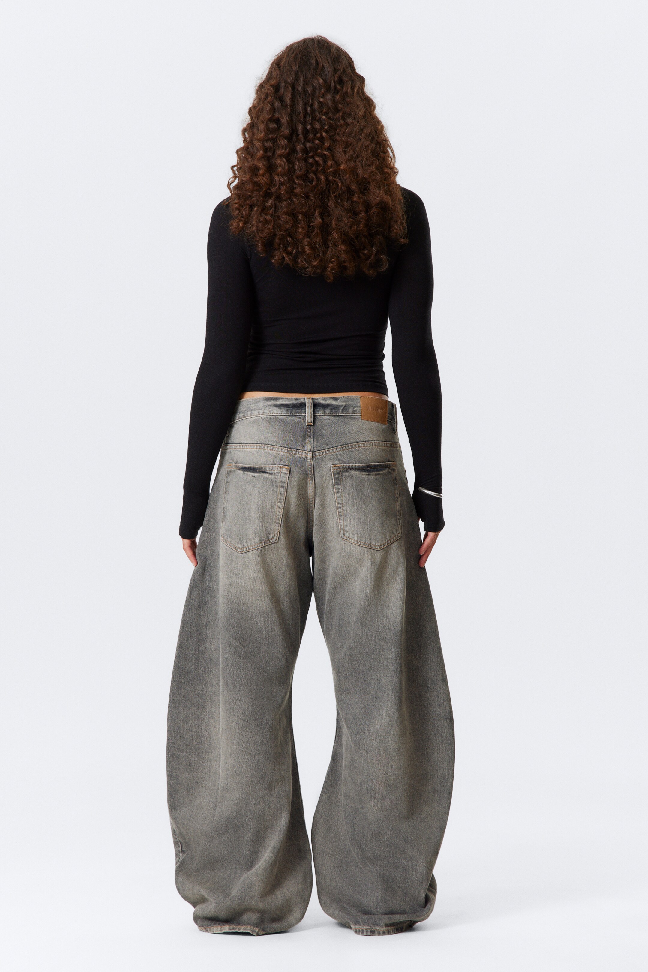 Agrandir l'image: Un pantalon large en jean délavé couleur charbon avec une coupe ample et des ourlets qui touchent le sol est présenté de dos, avec une ceinture visible dotée de passants et des poches arrière standard, ainsi qu'une étiquette de marque marron clair sur le côté droit. Un haut noir à manches longues avec des trous pour les pouces est porté, révélant partiellement le haut de la ceinture du jean.