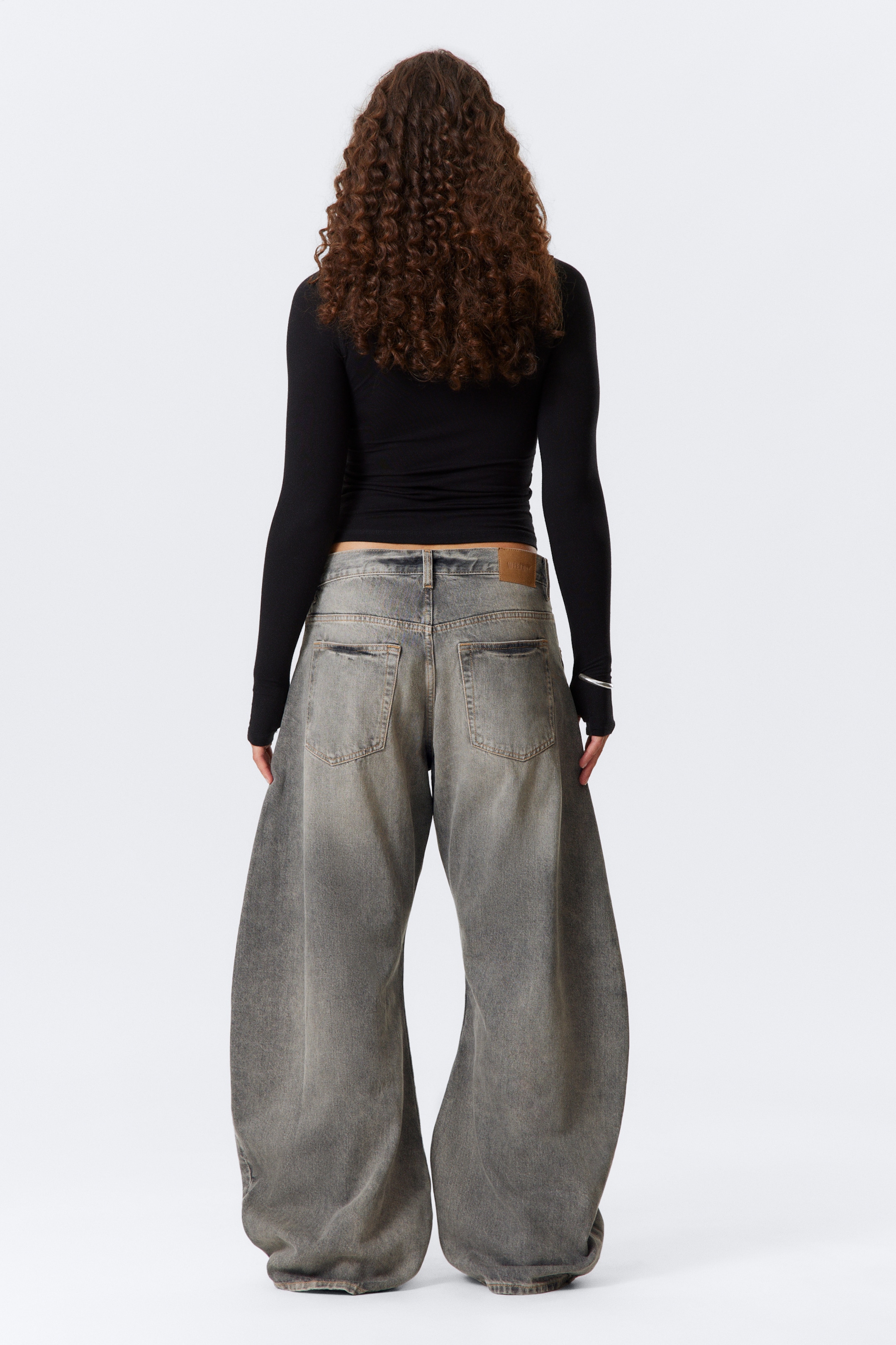 View larger image: A pair of faded charcoal wide-leg jeans with a baggy fit and puddle hems is being presented from the back, featuring a visible waistband with belt loops and standard back pockets, along with a light brown brand patch on the right side. A black long-sleeve top with thumbholes is being worn, partially revealing the top of the jeans' waistband.