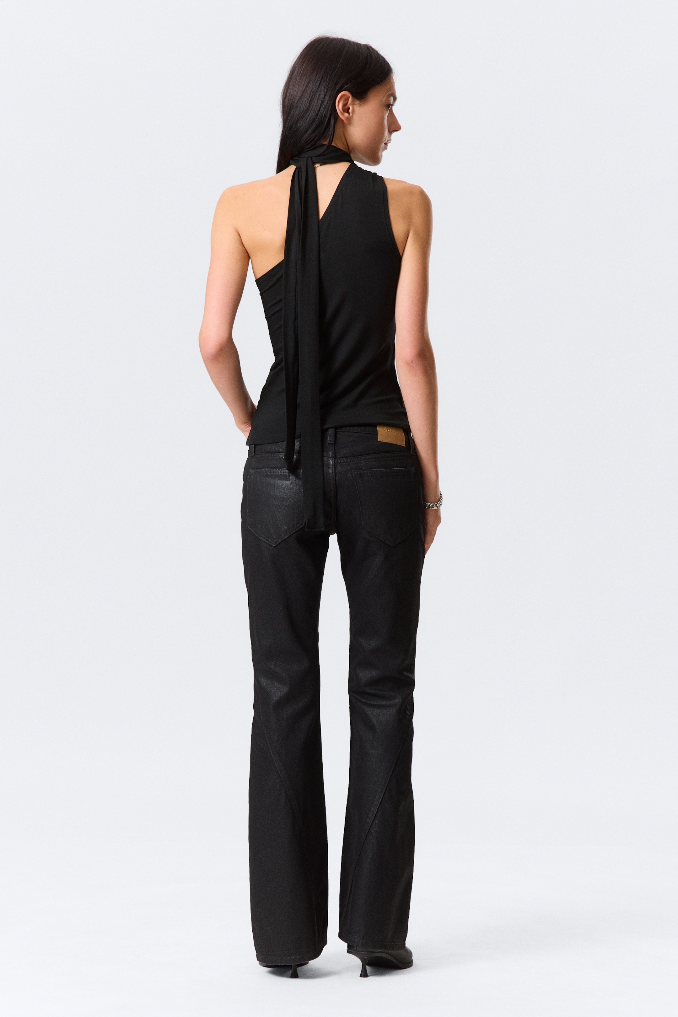 Grotere afbeelding bekijken: Een zwarte mouwloze top heeft een asymmetrisch ontwerp aan de achterkant met een V-uitsnijding, waaruit een lange, smalle strikdetail naar beneden hangt. Zwarte, gecoate-effect broek toont een uitlopende pijp met een opvallende V-vormige naad op de onderrug van elke pijp, en een bruine leren patch is zichtbaar op de achterste tailleband.