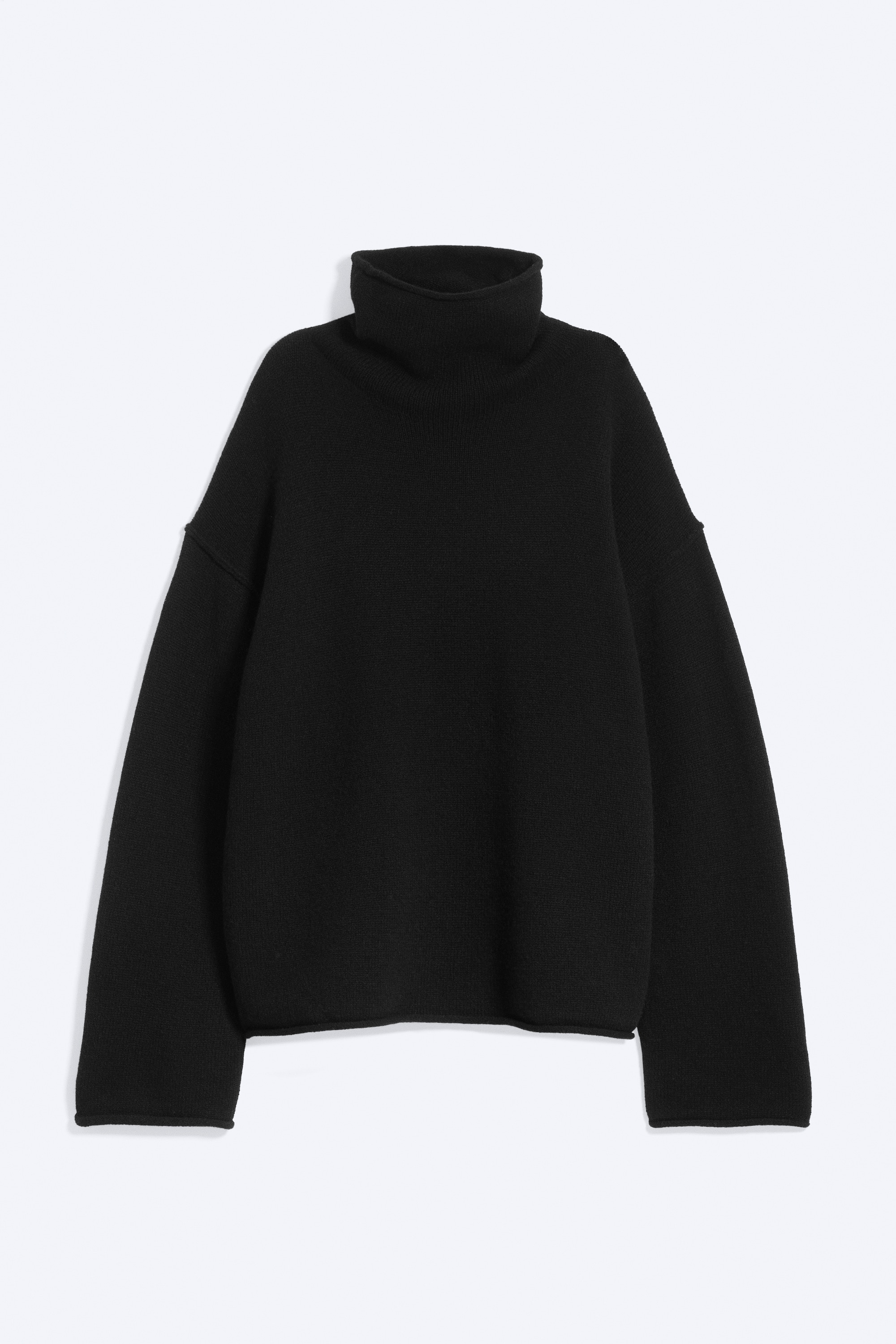 Un maglione a collo alto in lana nera dalla vestibilitÃ  morbida, con collo alto a imbuto e spalle scese, indossato dalla modella. Il maglione Ã¨ abbinato a pantaloni cargo mimetici verdi e stivali neri, mentre la modella tiene in mano una piccola borsa in tela beige.