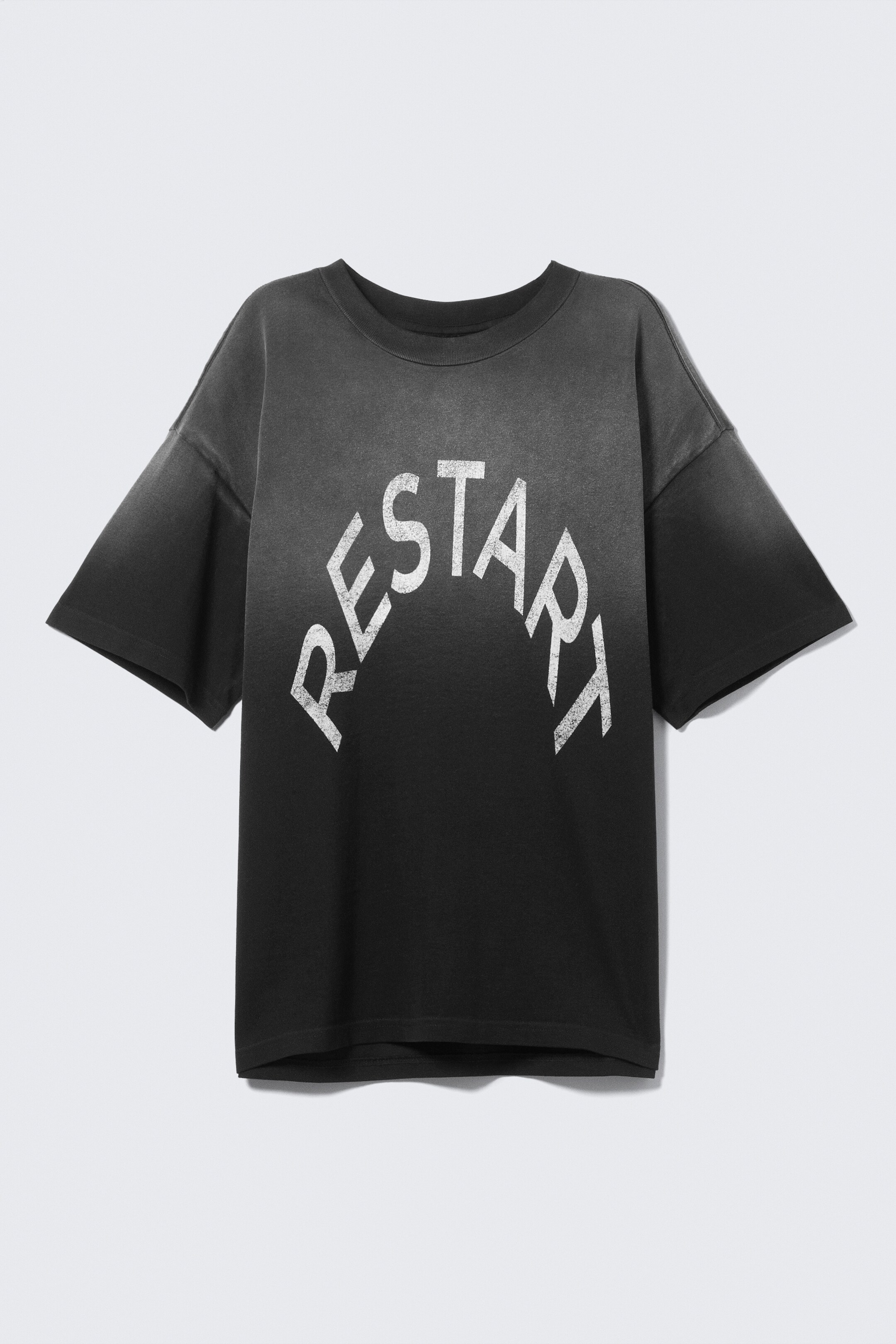 Visa större bild: En löst sittande, svart t-shirt är vänd FRAMÅT och visar en urtvättad effekt som bleknar från svart till en ljusare grå vid toppen och ärmarna. Den har en boxig passform med nedhasade axlar, en rundad hals och dubbelsydda fållar, medan en sliten vit "RESTART"-grafik böjer sig över bröstet.
