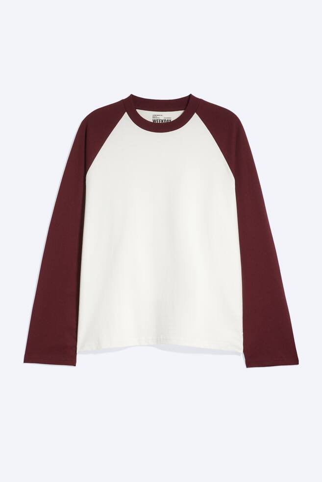 A long-sleeved raglan T-shirt is shown from the front, featuring a relaxed, boxy fit with an off-white body and contrasting burgundy sleeves. The T-shirt showcases a matching burgundy ribbed crew neckline and straight-cut hems. - 1