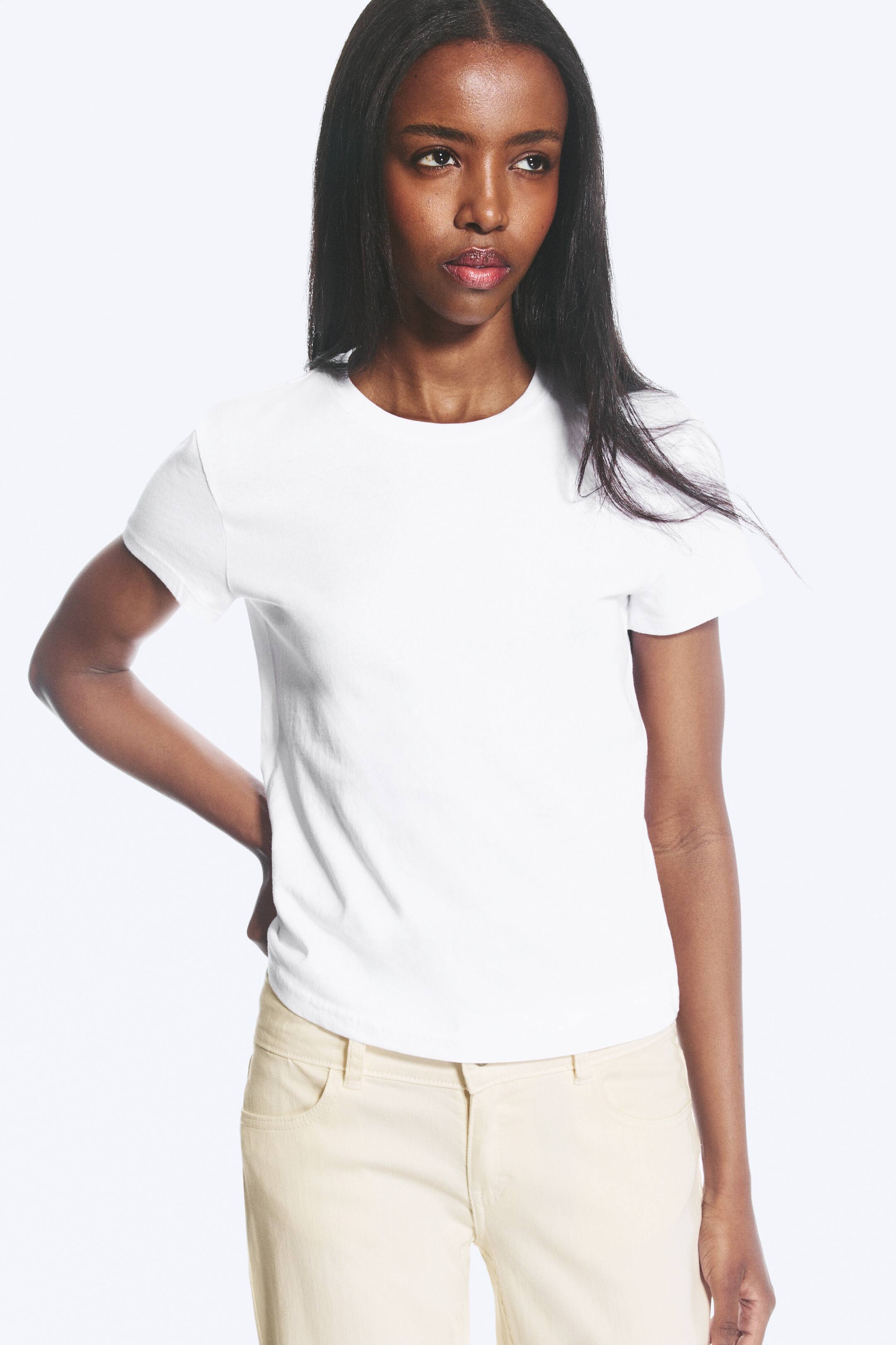 Grotere afbeelding bekijken: Een wit T-shirt met ronde hals en korte mouwen in een slanke pasvorm wordt gedragen door het model. Het T-shirt is gestyled met lichtgekleurde broek, en het model plaatst haar linkerhand op haar heup.