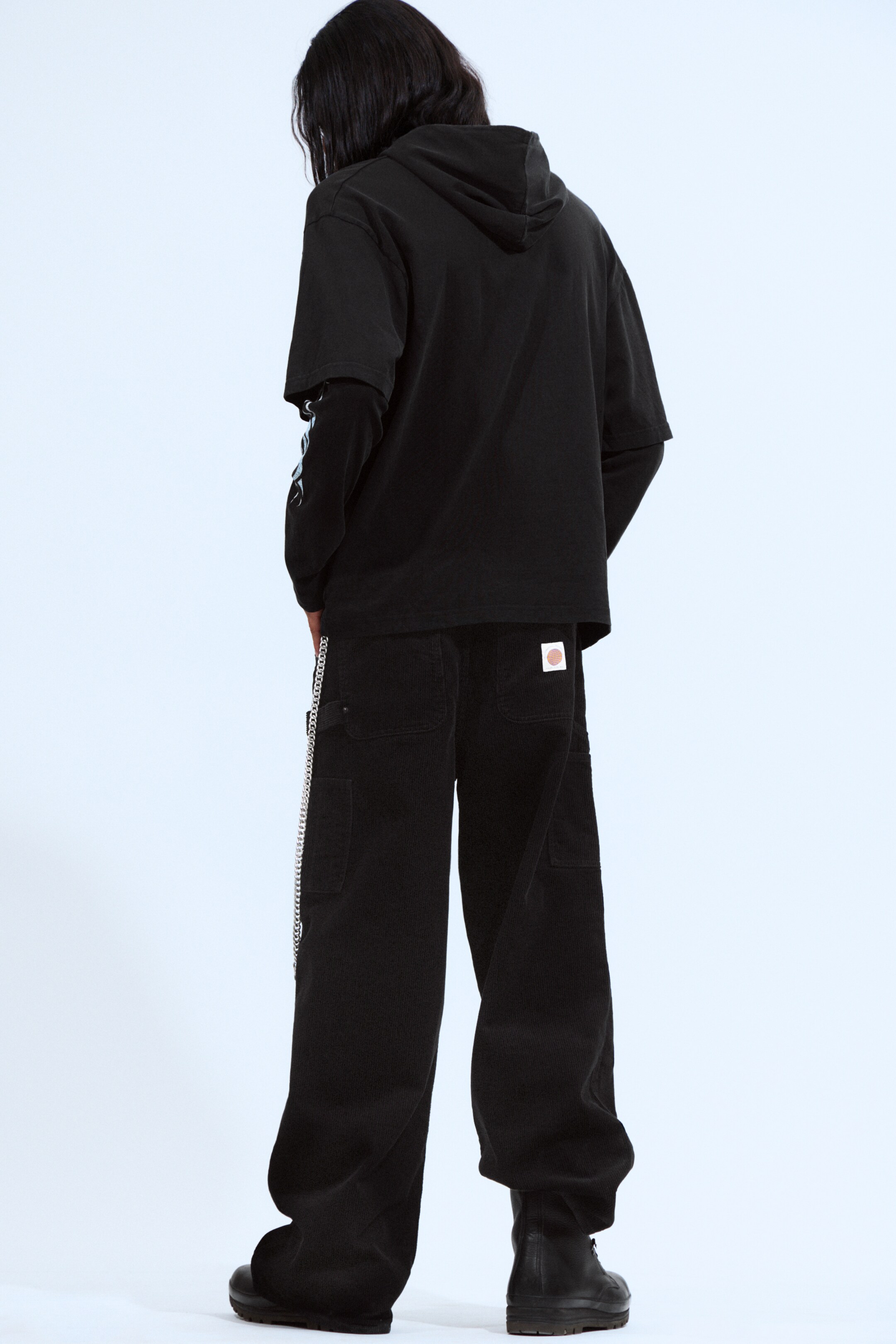 View larger image: A black short-sleeved hoodie is being worn over a black long-sleeved shirt, which is featuring light-colored graphics on its sleeves, seen from a back view. Black wide-leg corduroy pants are also being worn, displaying a silver-toned chain on the left side and a light-colored patch on the right back pocket.