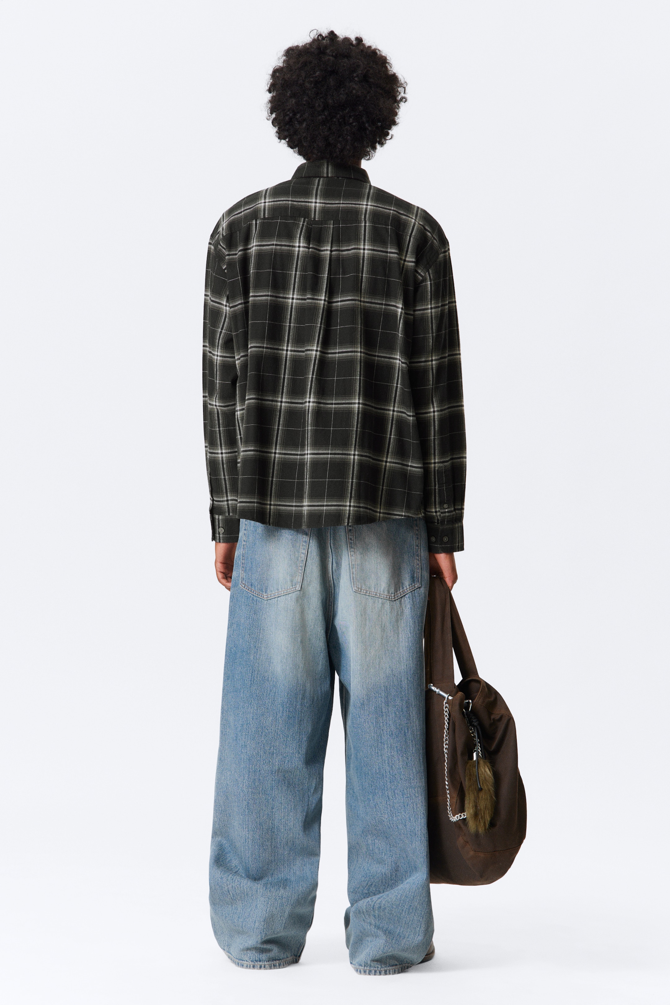Grösseres Bild ansehen: Ein dunkles Karohemd, von hinten gesehen, wird mit hellblauen, weit geschnittenen, ausgewaschenen Jeans mit sichtbaren Gesäßtaschen getragen. Eine braune Tragetasche, verziert mit einer silbernen Kette und einem flauschigen Anhänger, wird getragen.