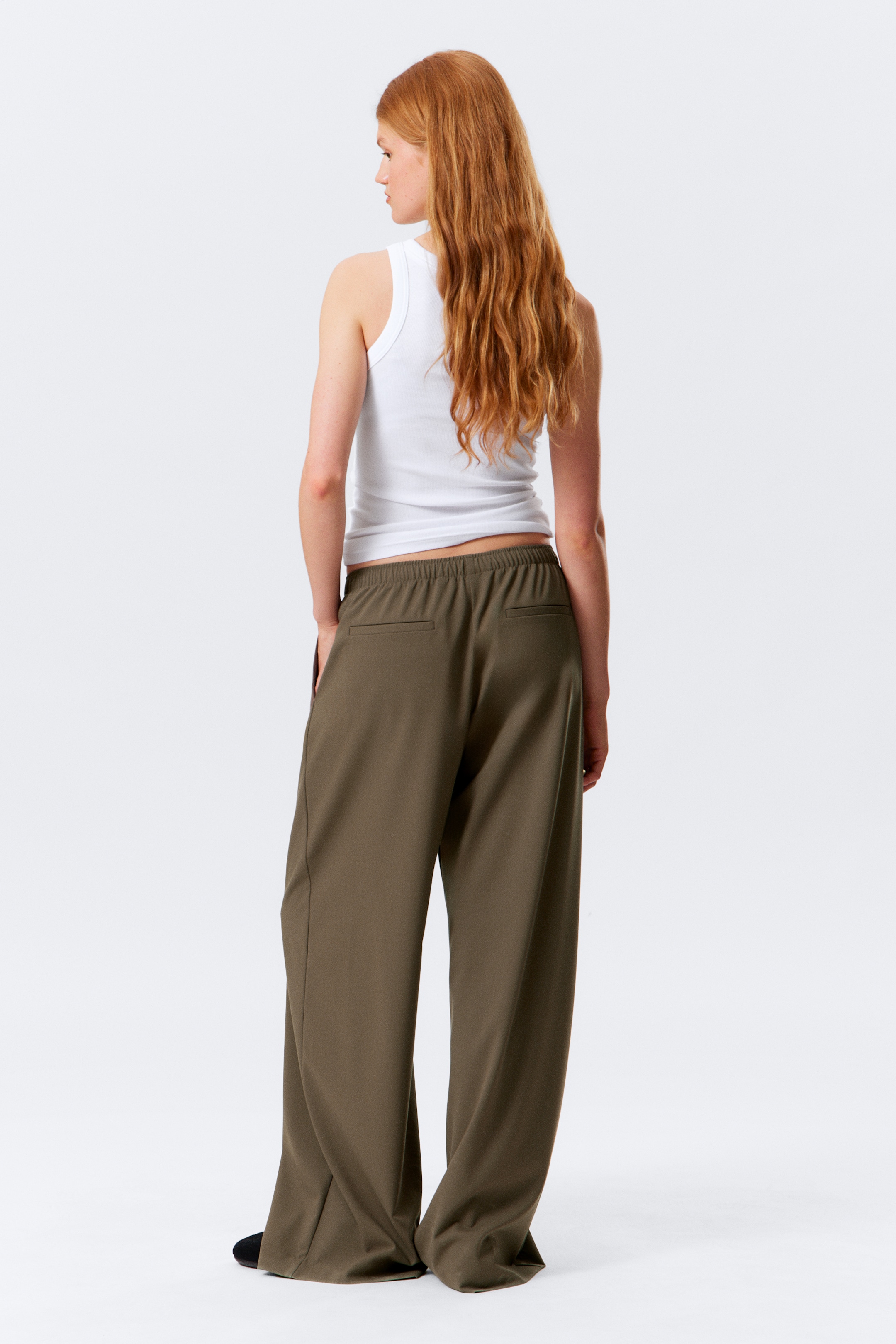 Grösseres Bild ansehen: Olivgrüne Hosen mit weitem Bein, die über einen elastischen Bund und zwei rückseitige Leistentaschen verfügen, werden von hinten präsentiert. Ein weißes geripptes Tanktop wird über den Hosen getragen.