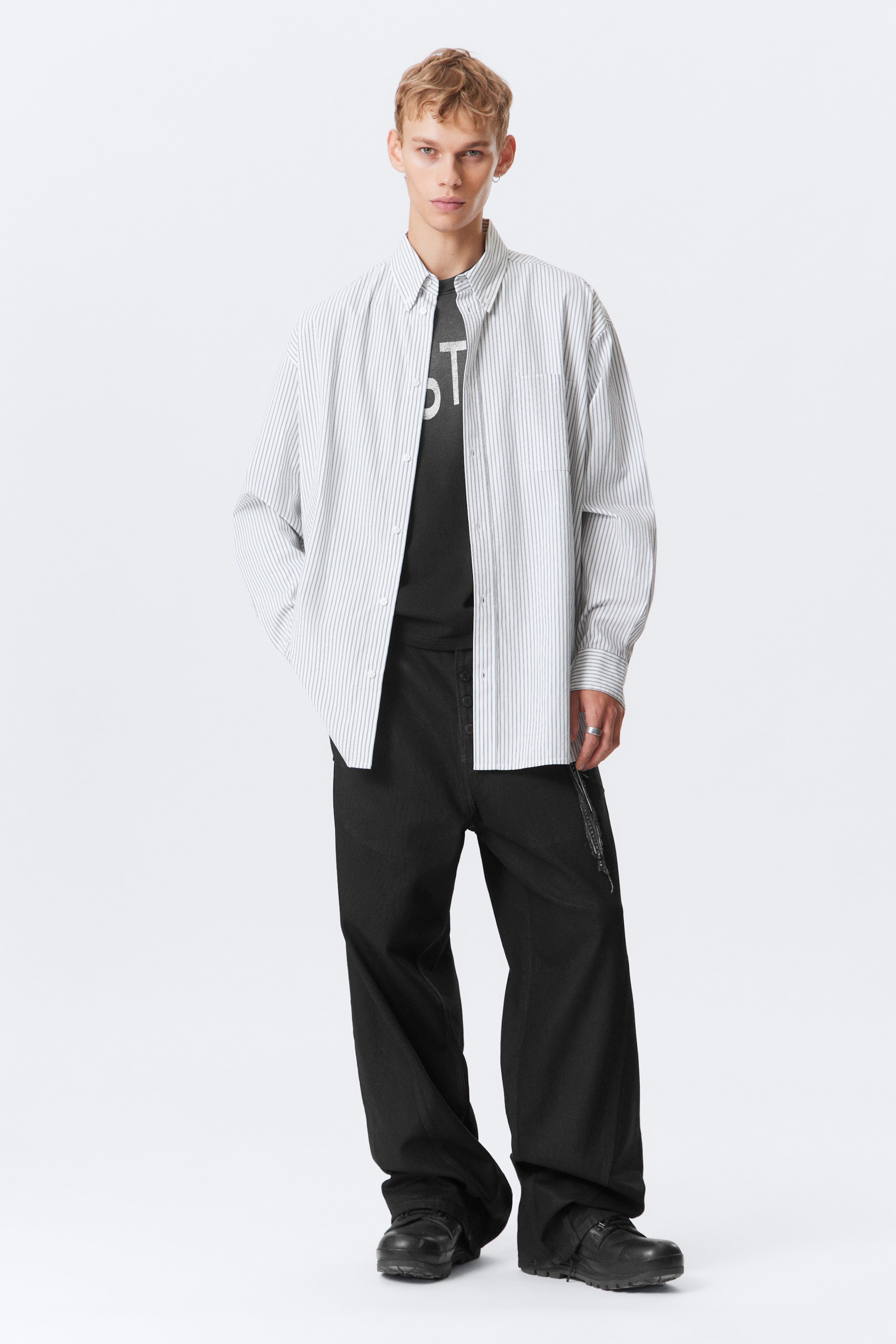 View larger image: An oversized white button-down shirt, featuring thin vertical dark stripes and a button-down collar, is being worn open over a dark crew-neck t-shirt with a partial white graphic. Black wide-leg pants with a visible button fly are being worn, creating a loose fit that pools at the ankles.