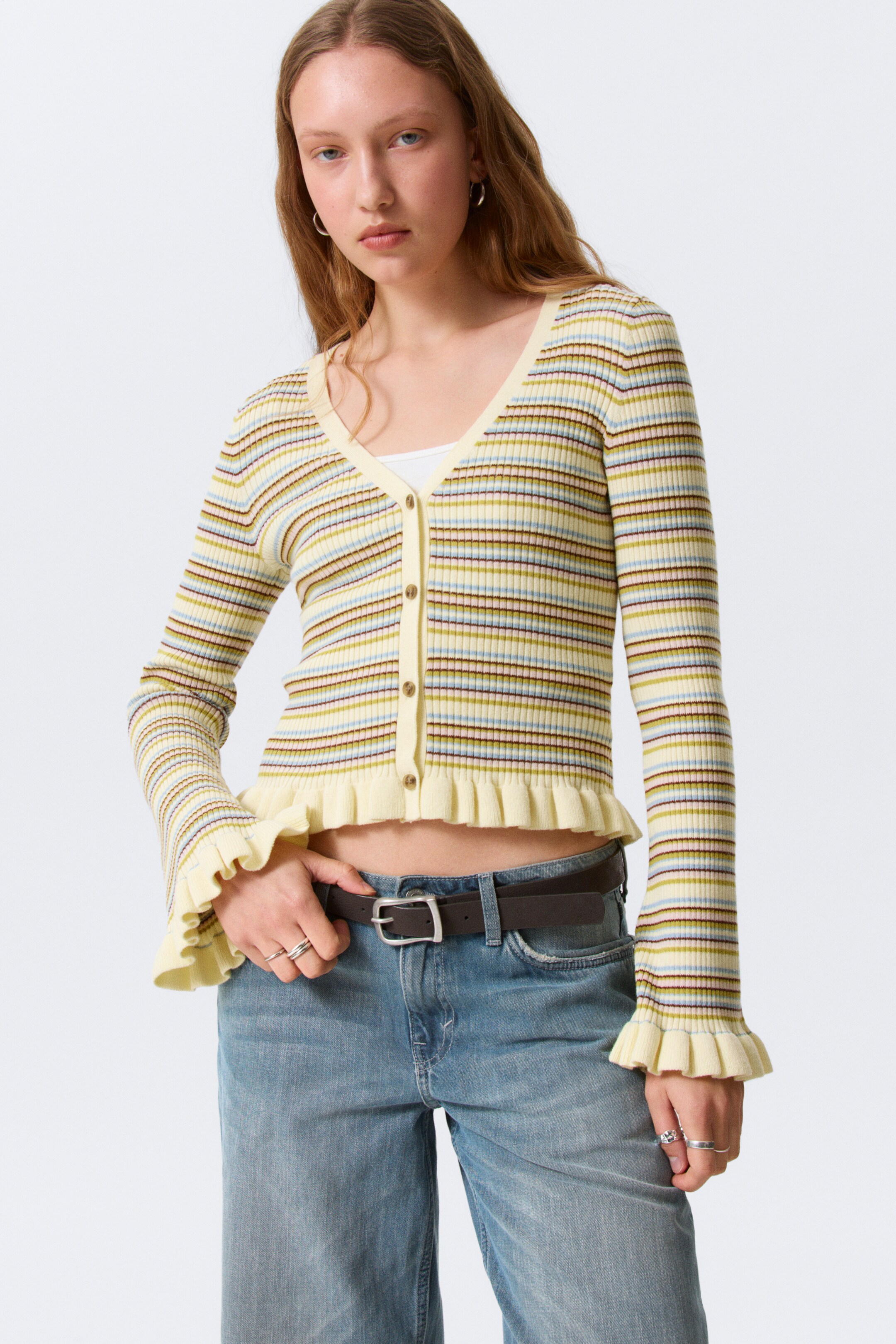 Преглед на по-голямо изображение: Рипсено плетено жилетка с V-образно деколте и предно закопчаване с копчета в светъл цвят. Представя се с хоризонтални ивици в жълто, светло синьо, маслинено зелено и кафяво, и е проектирана с дълги ръкави, маншети с къдрици и къс подгъв с къдрици, като под нея се вижда бяла горна част.