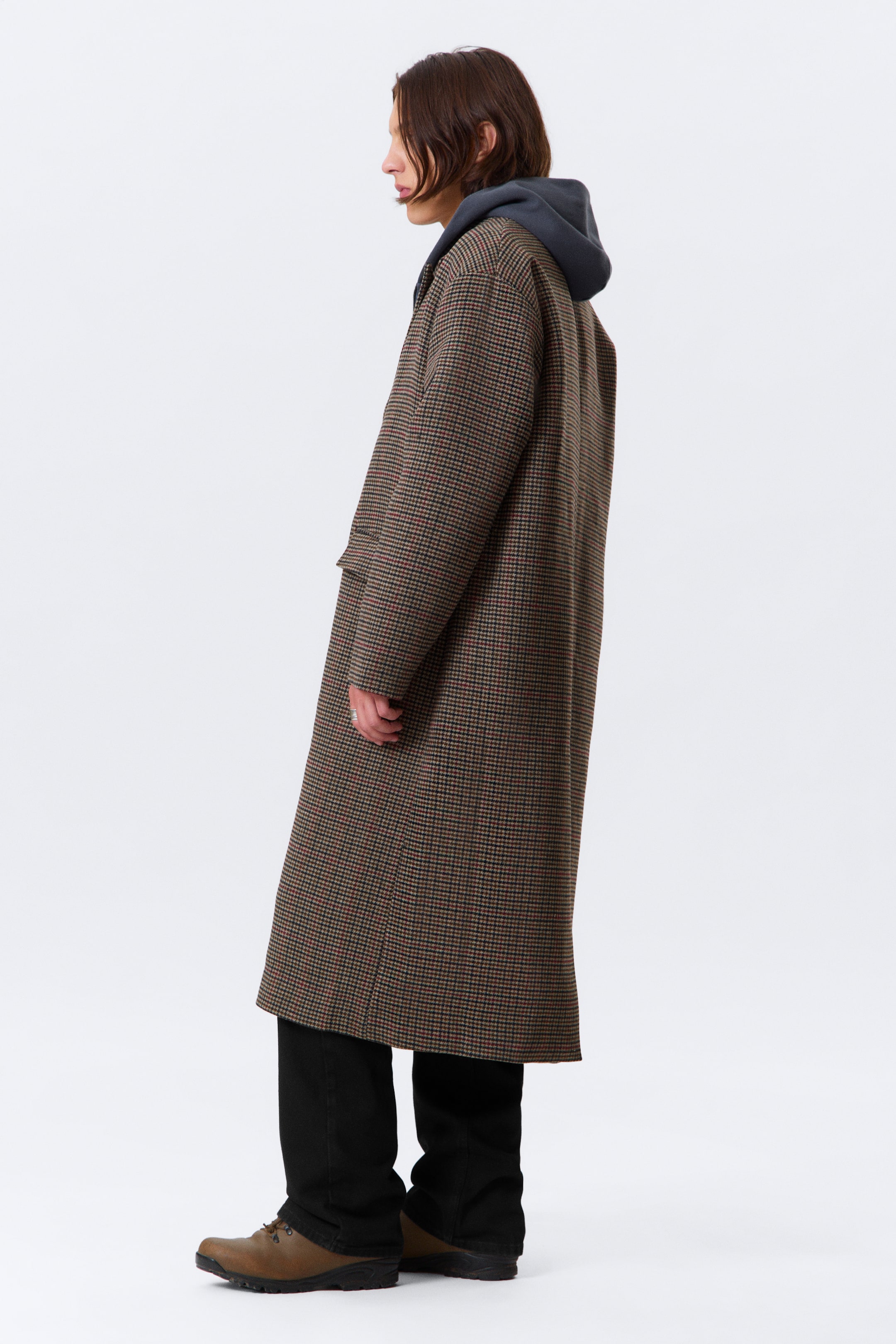 Visualizza immagine più grande: Un lungo cappotto ampio in un motivo a micro quadri marrone e nero con sottili accenti rossi viene presentato da un'angolazione laterale, mostrando la sua lunghezza completa e la silhouette rilassata. Presenta una patta della tasca visibile ed è stratificato sopra una felpa con cappuccio grigio scuro.